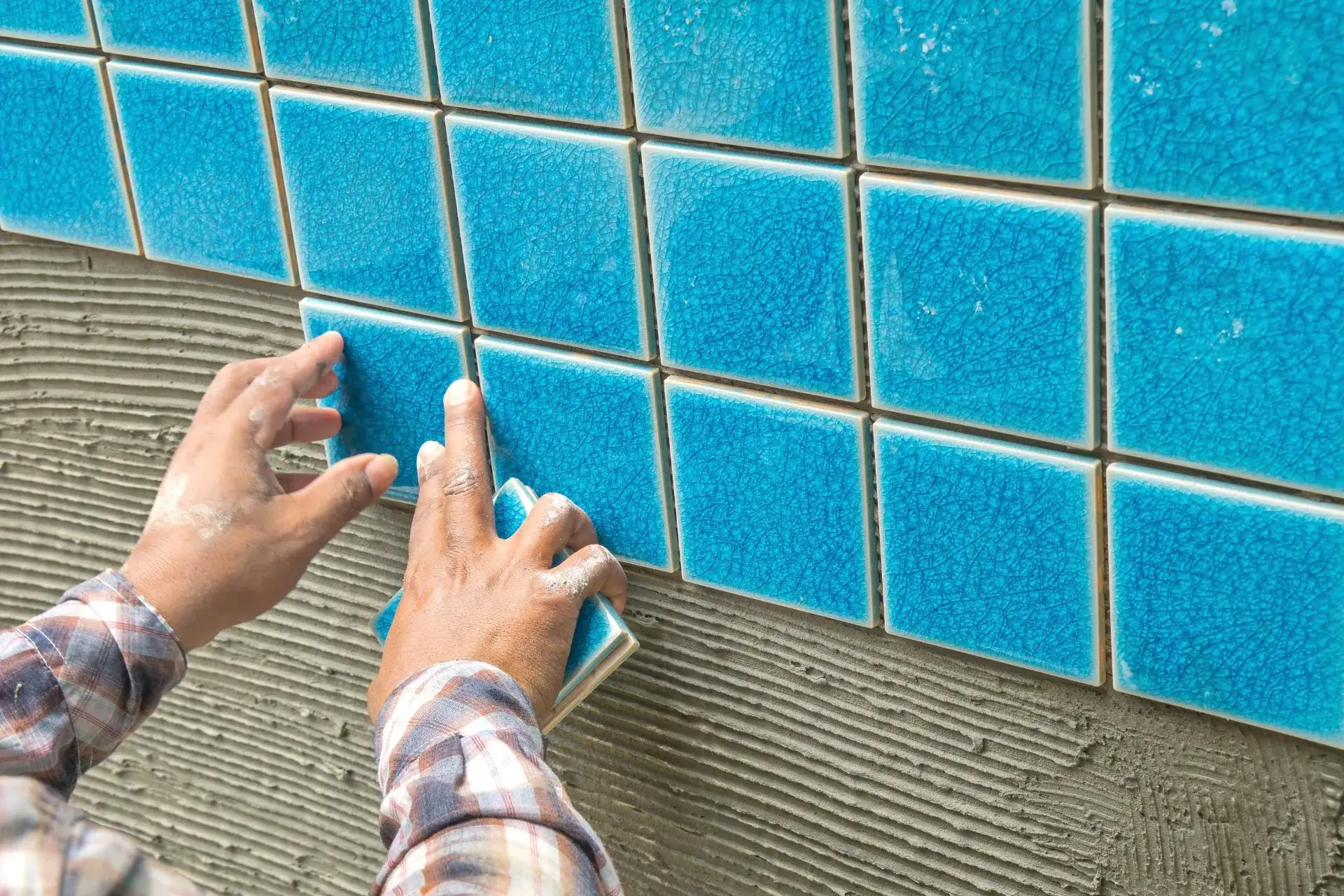 Hands installing blue ceramic tiles on wall over grey adhesive.