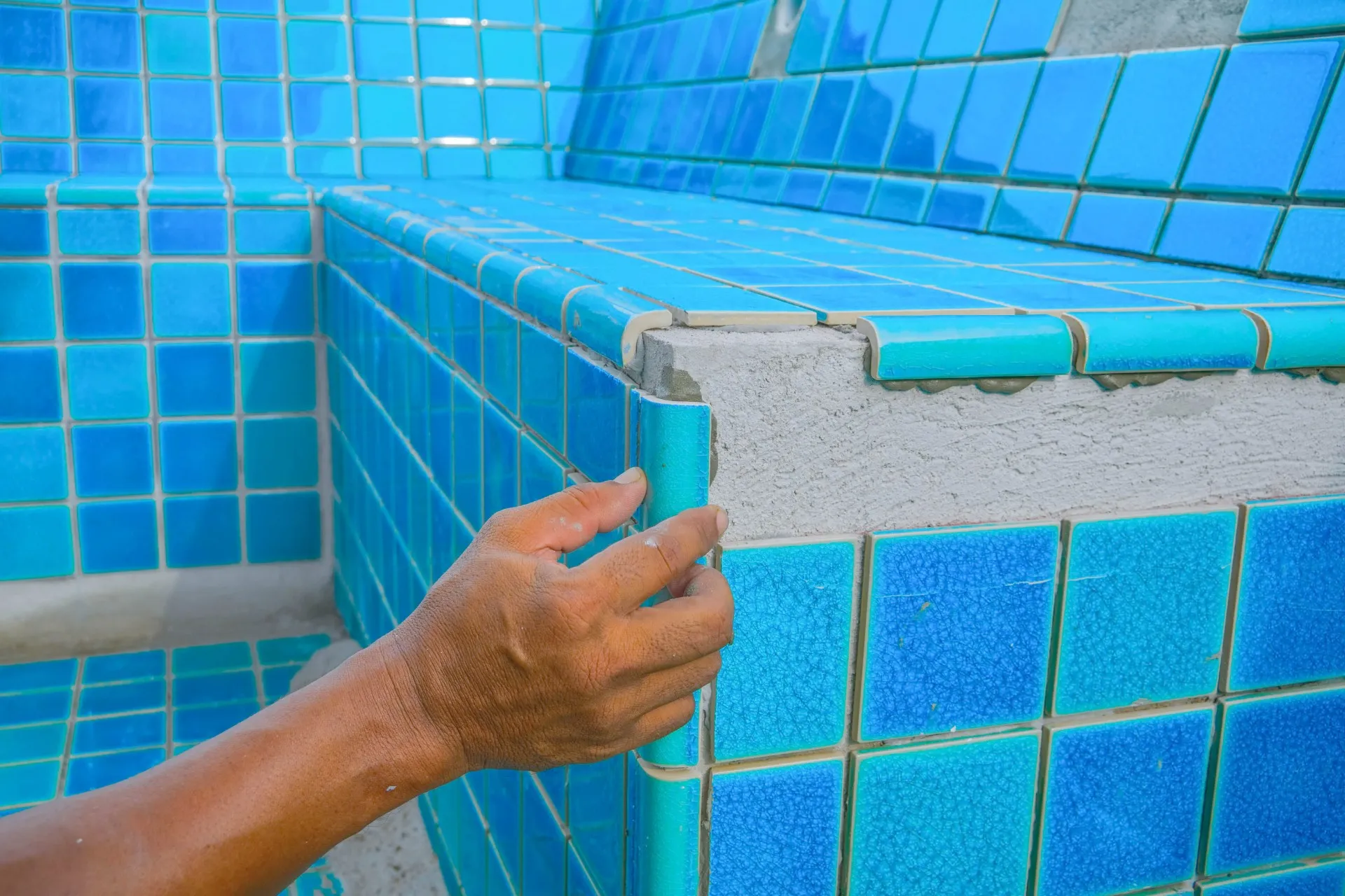 Hand holding a broken blue ceramic corner tile on a tiled pool edge under repair.