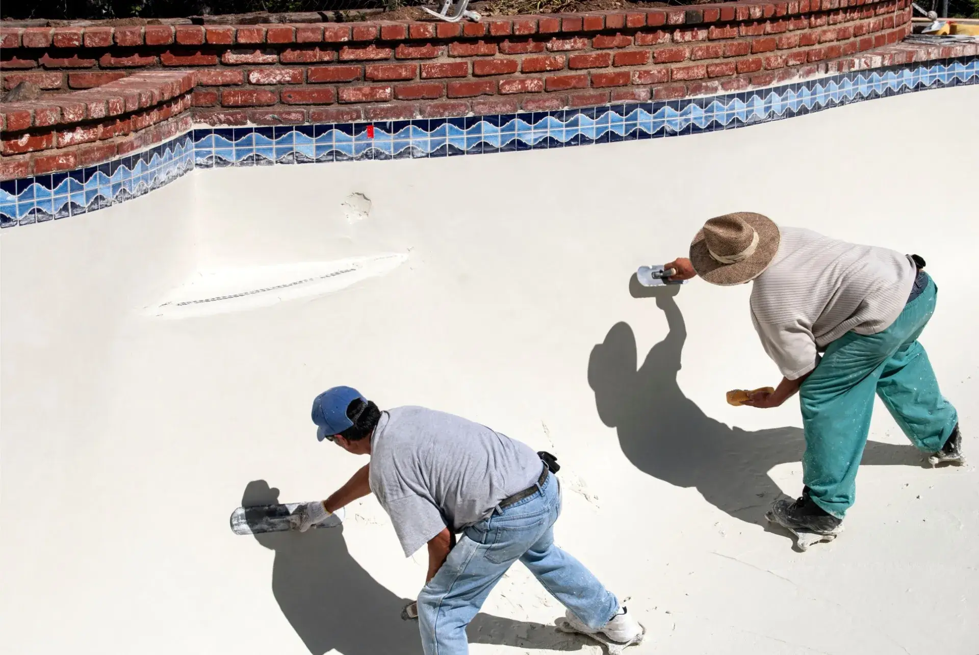Two workers smoothing the surface of an empty swimming pool during repair work.