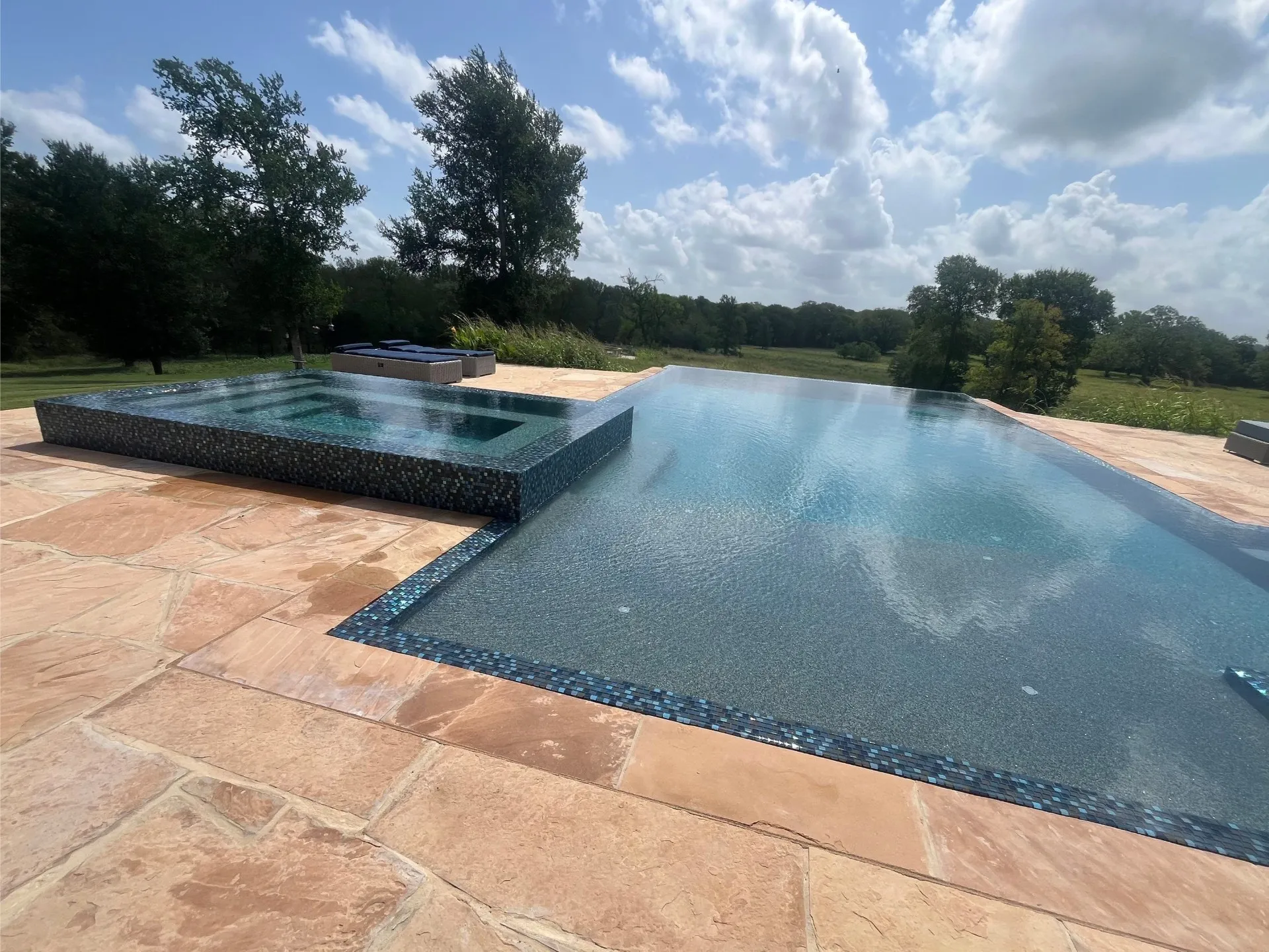 Infinity edge swimming pool with an adjacent raised hot tub surrounded by tan stone tiles and trees in the background.