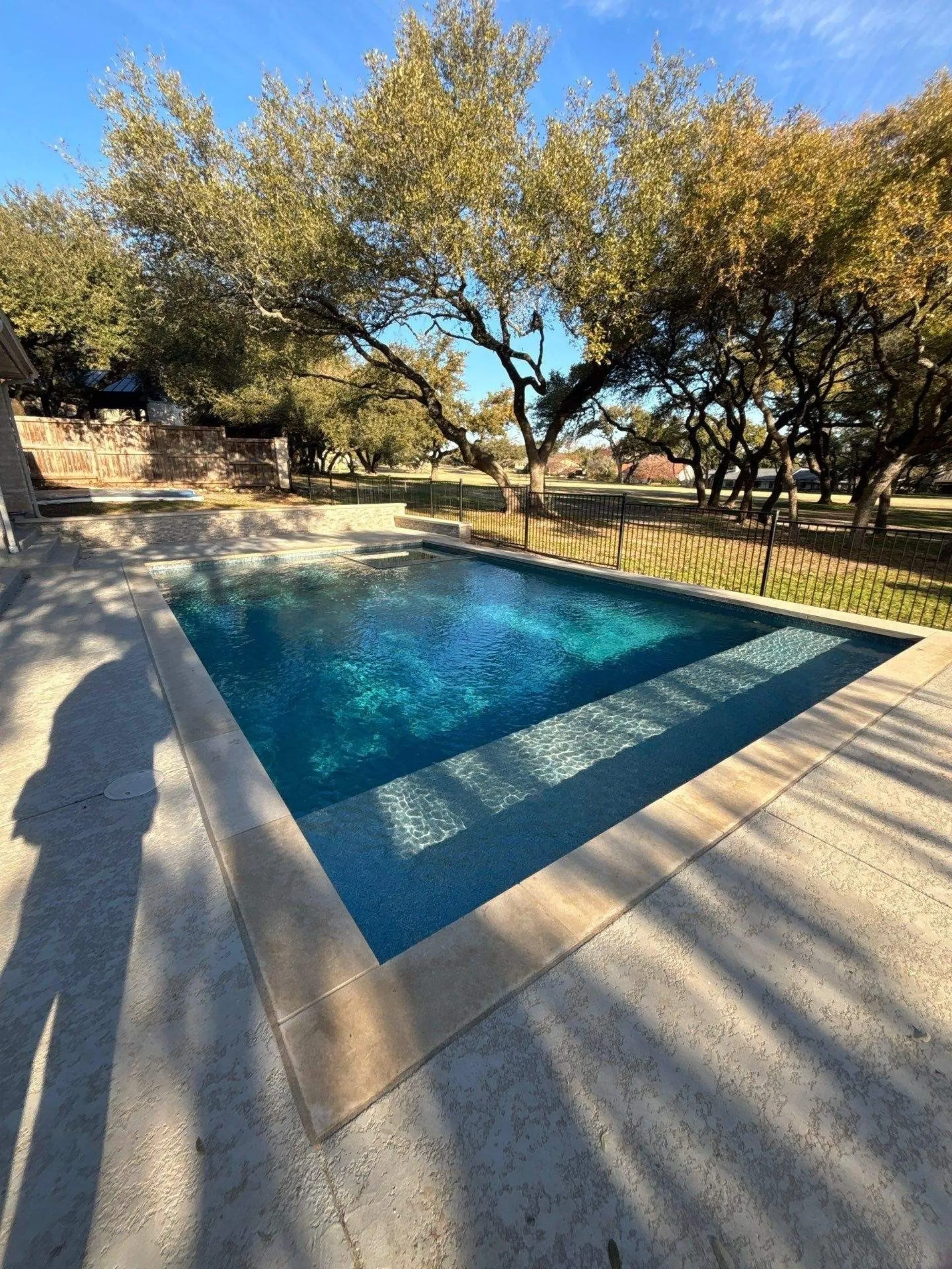 Rectangular backyard swimming pool with clear blue water surrounded by concrete decking and trees in the background.