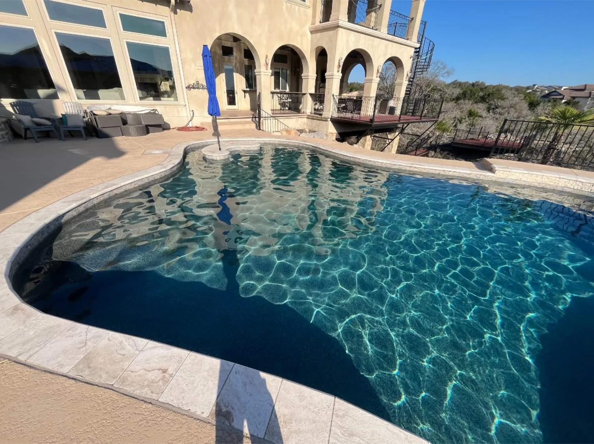 Clear blue swimming pool surrounded by beige tile decking with a beige stucco house and patio furniture in the background.