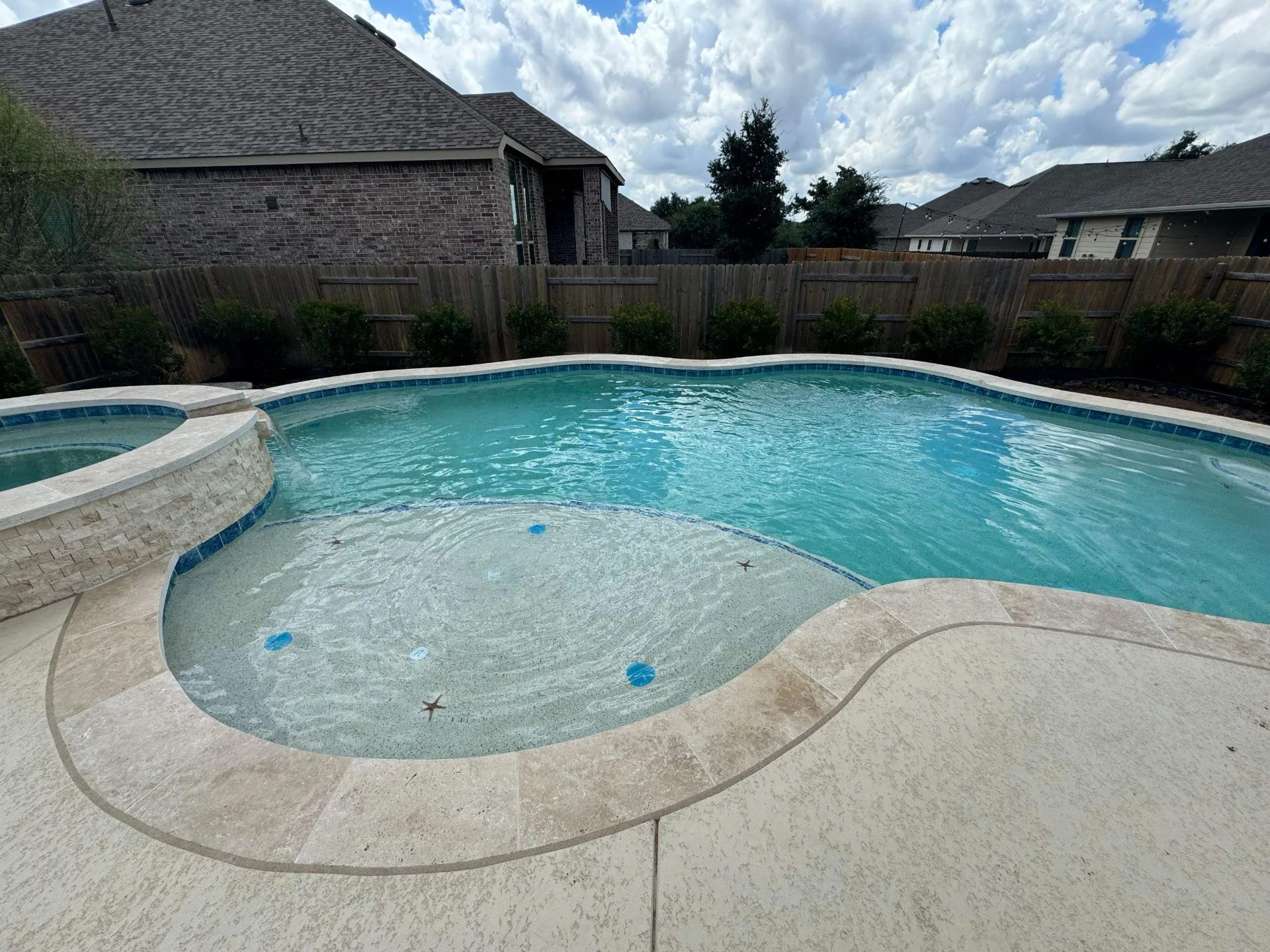 Backyard swimming pool with a shallow tanning ledge and attached spa, surrounded by a stone deck and wooden fence.