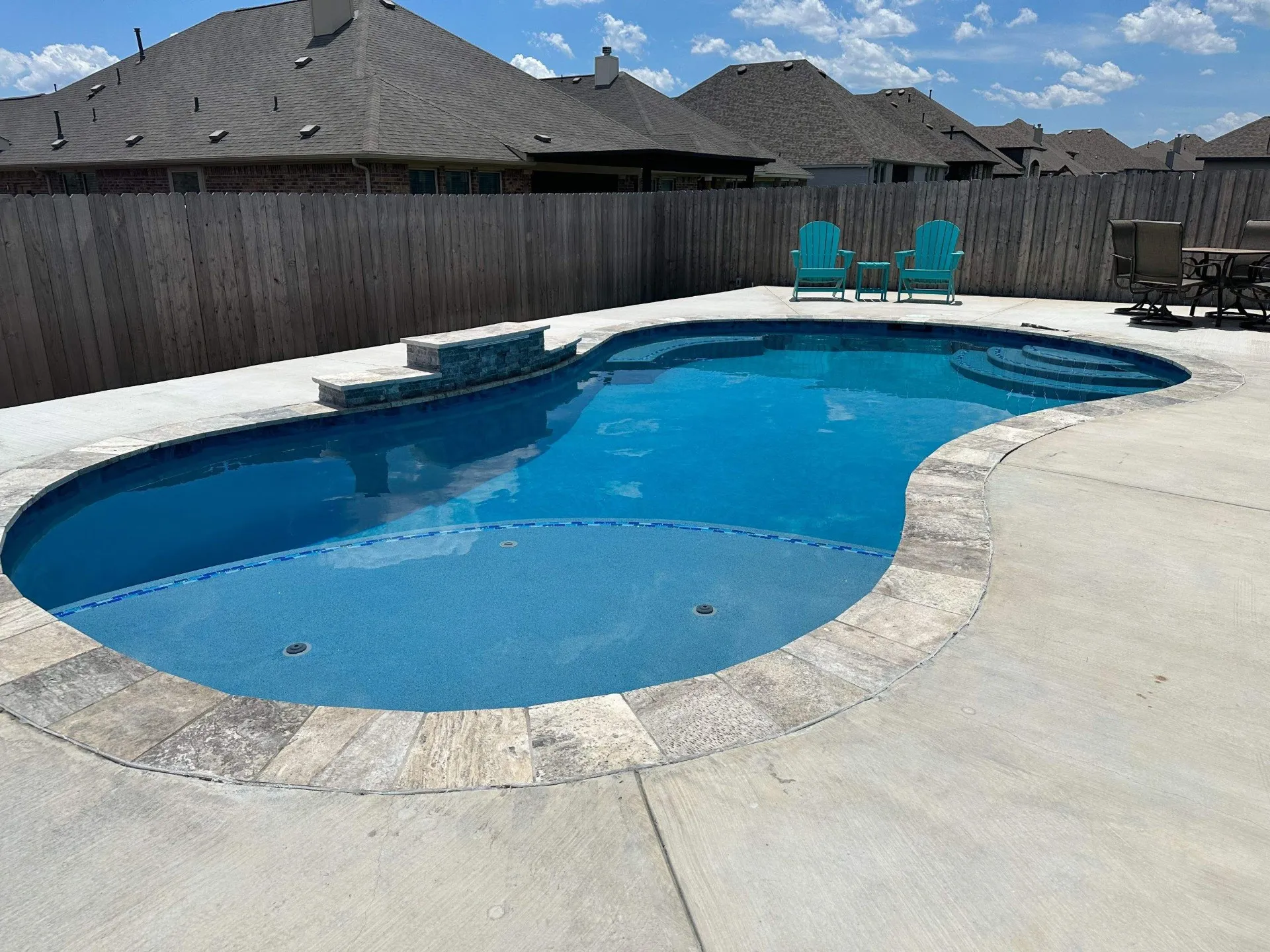 Backyard swimming pool with stone edging, two turquoise chairs and small table, and a high wooden fence under a partly cloudy sky.