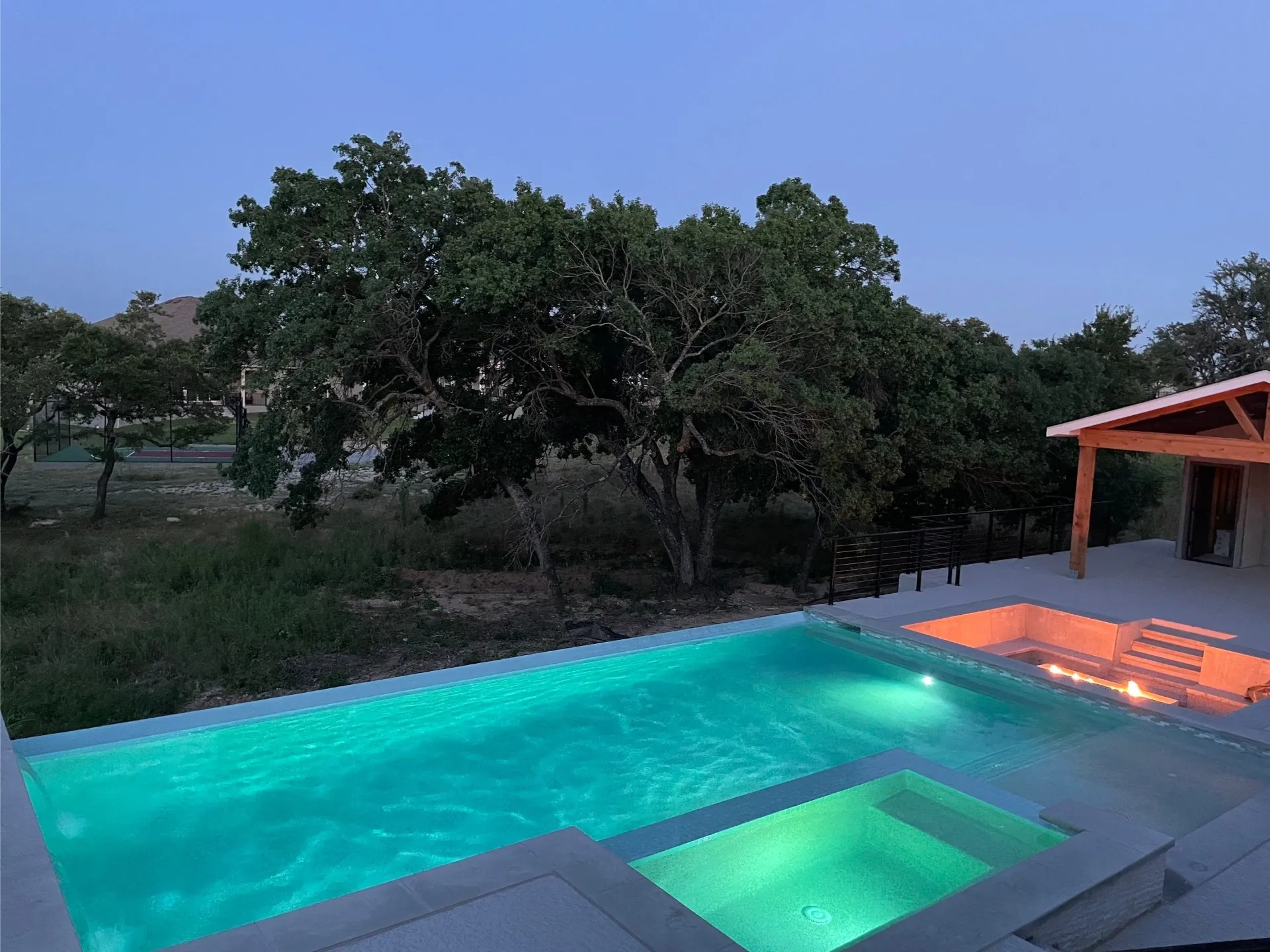 Outdoor swimming pool and hot tub illuminated with blue-green lights at dusk, with trees and a small pavilion nearby.