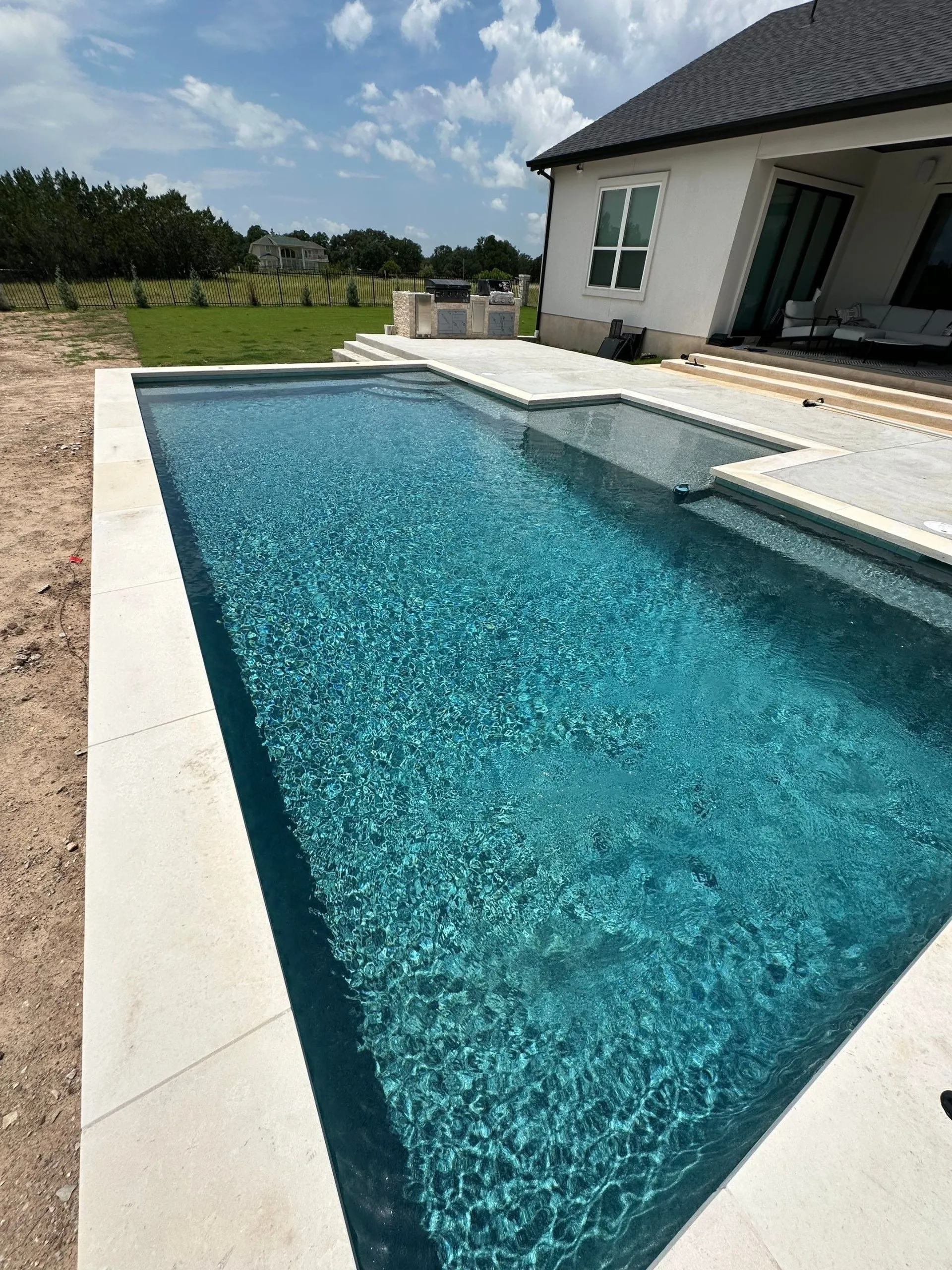 Modern rectangular swimming pool with clear blue water next to a white house patio under a partly cloudy sky.