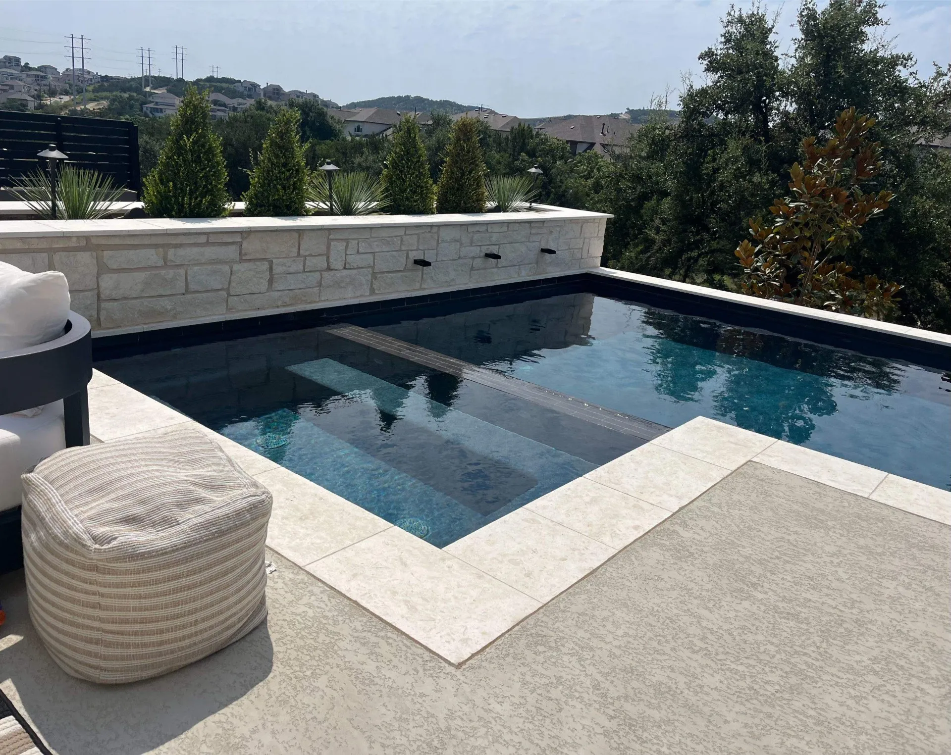 Modern outdoor pool with stone tile edging, built-in steps, and a planter wall with greenery, overlooking a hilly residential area.
