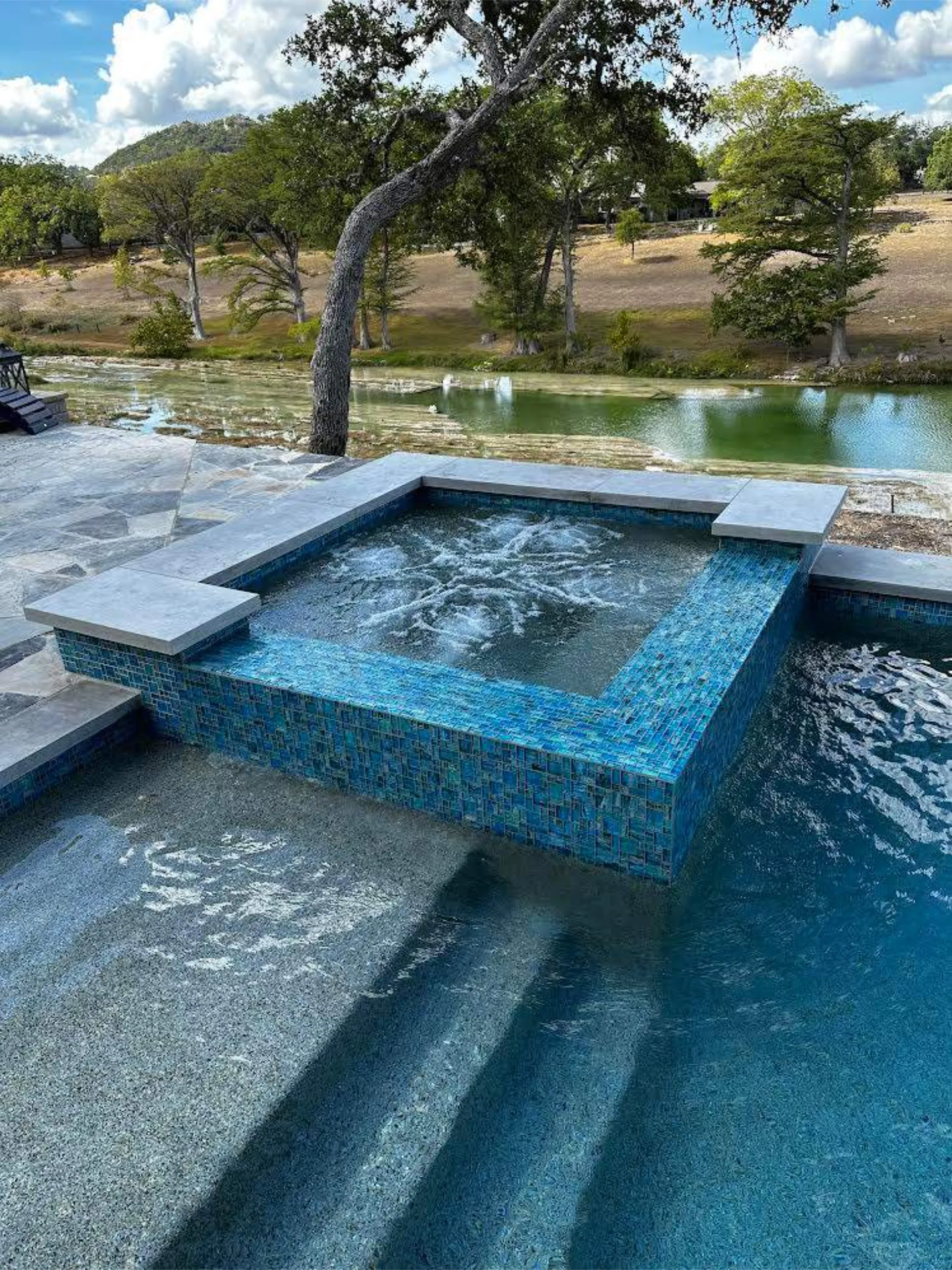 Outdoor pool area with a square blue-tiled hot tub bubbling next to a shallow pool and overlooking a green river and trees under a partly cloudy sky.