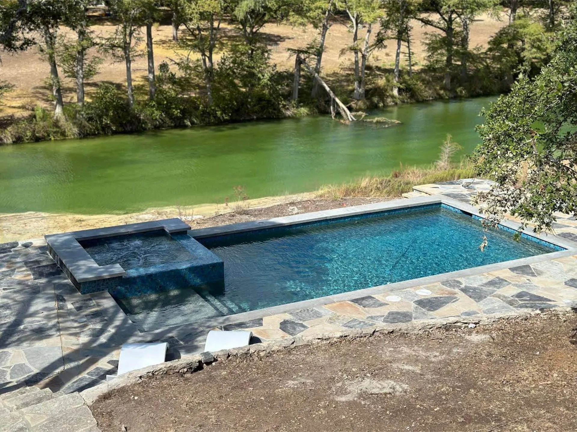 Rectangular stone-tiled pool with an attached square hot tub, overlooking a green river and trees.