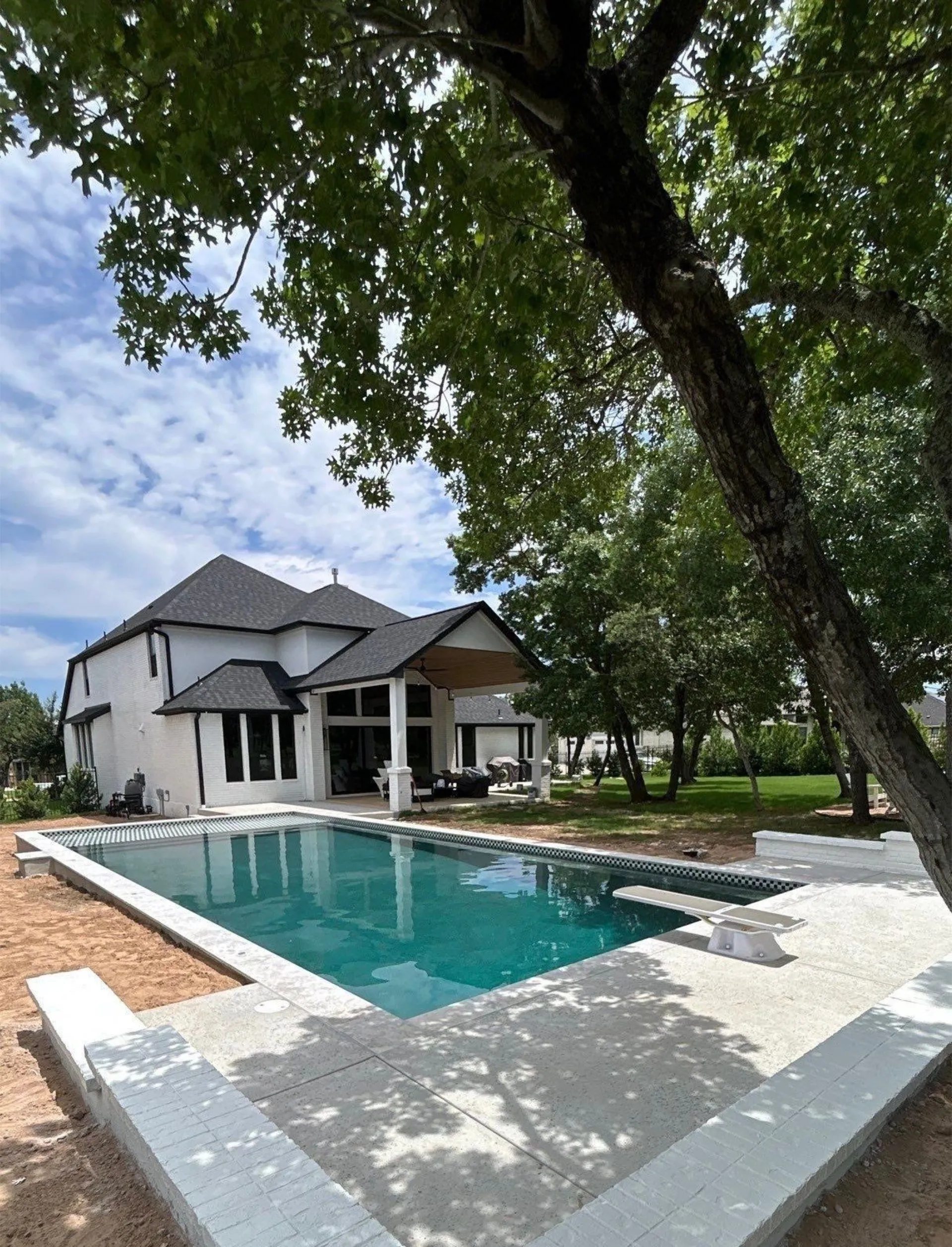 Modern white two-story house with a covered patio and a rectangular swimming pool surrounded by concrete decking and trees.