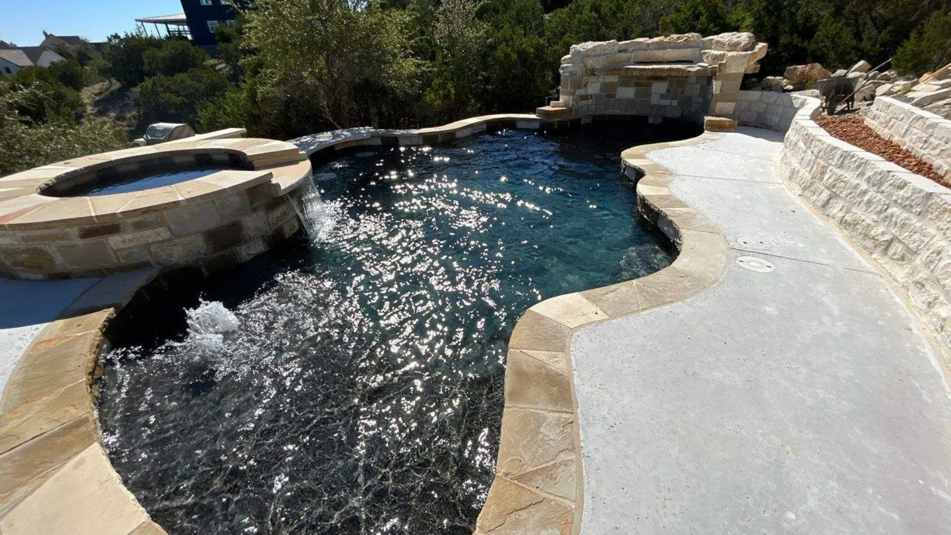 Outdoor swimming pool with a stone-bordered hot tub and waterfall feature, surrounded by a concrete deck and stone walls.