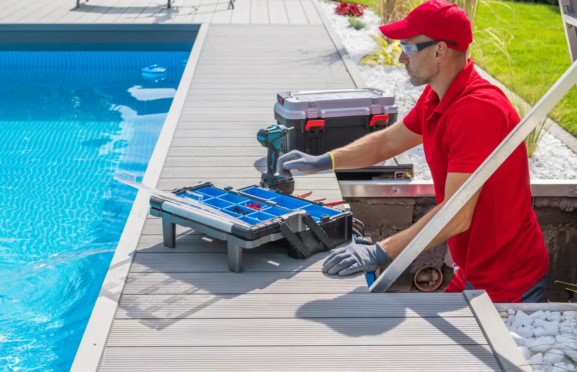 Technician in red uniform working by a swimming pool with a toolbox and electric drill.