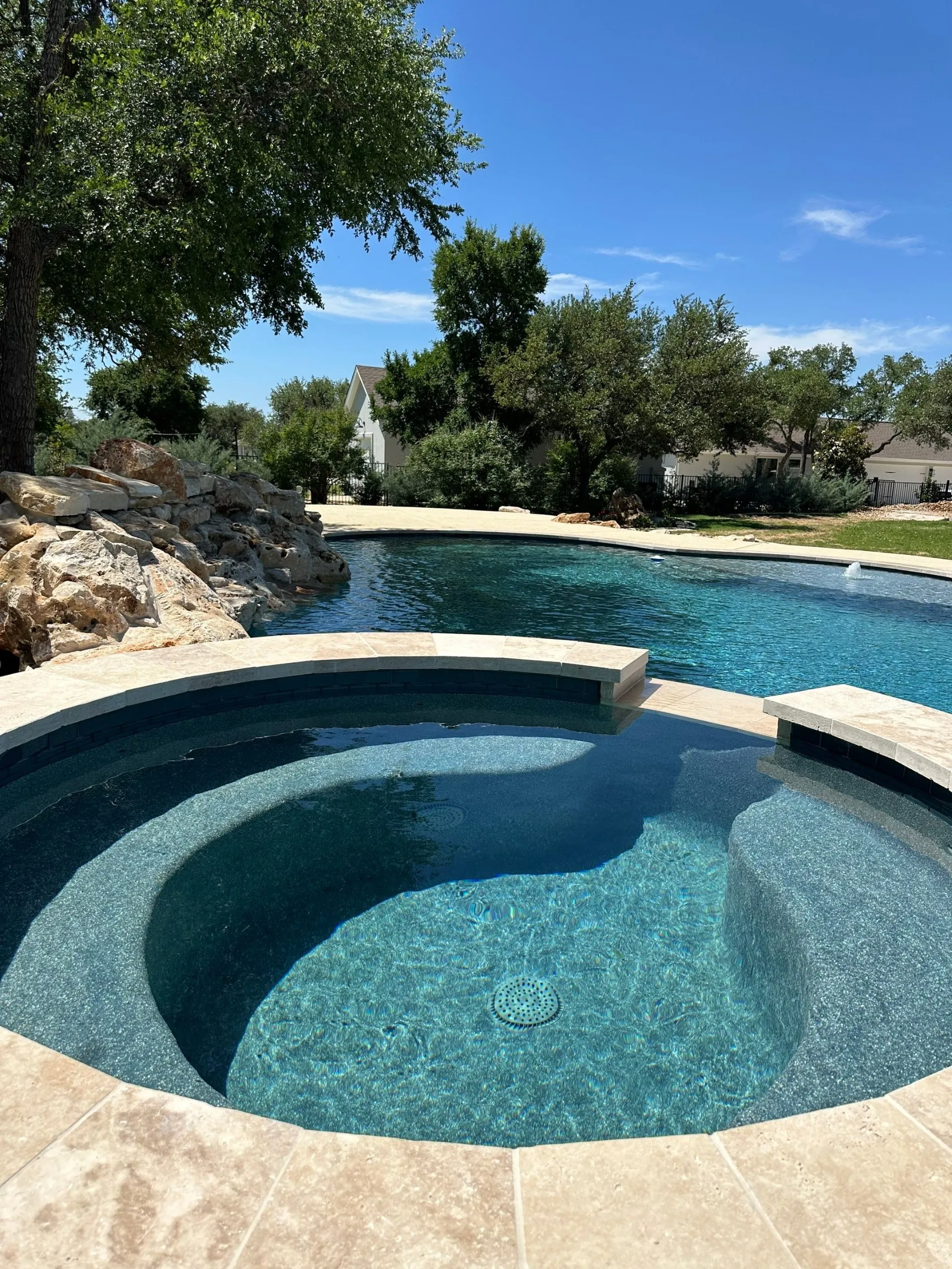 Clear blue circular hot tub adjacent to a large swimming pool with surrounding trees and a partly cloudy blue sky.