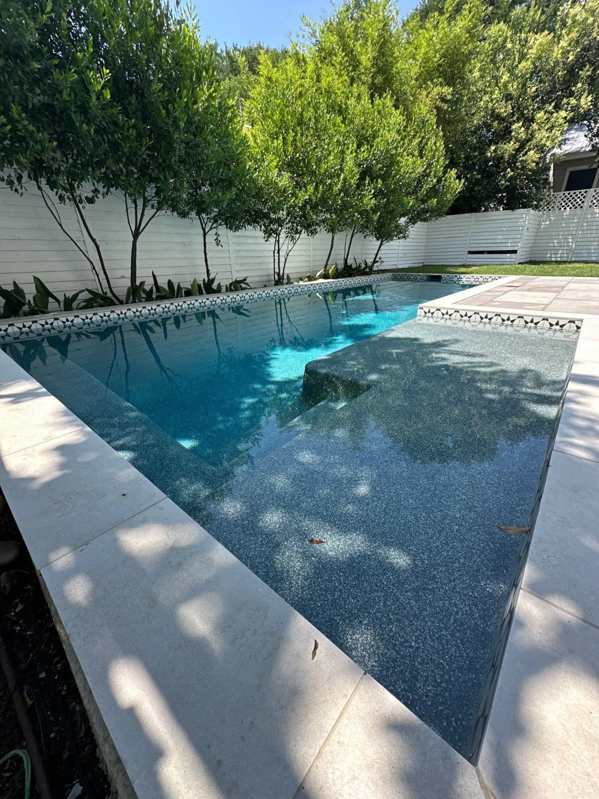 Modern rectangular outdoor swimming pool with clear blue water, surrounded by white tiled deck and green trees with white fence in the background.