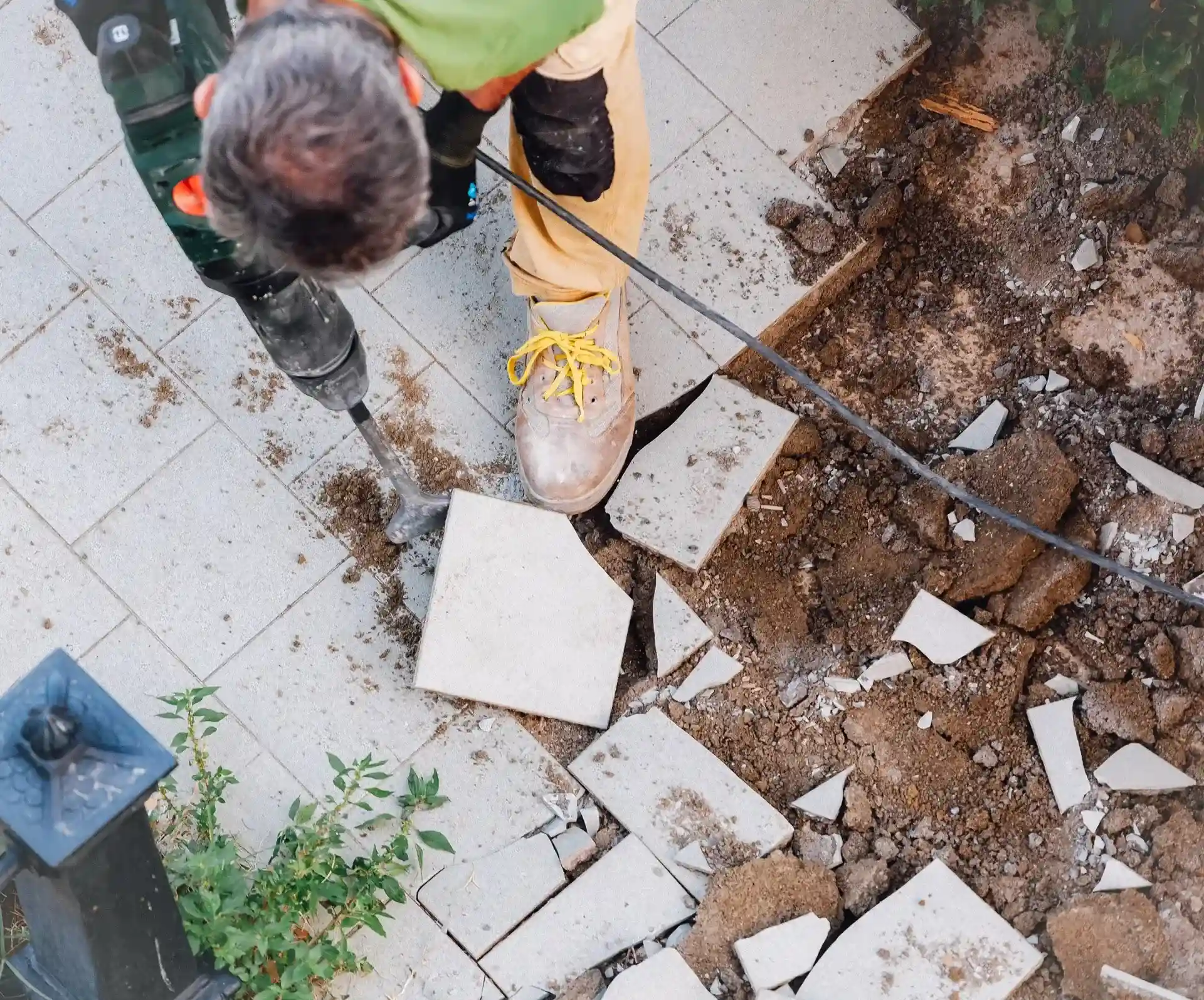 Construction worker using a power drill to break concrete tiles on the ground near soil and plants.