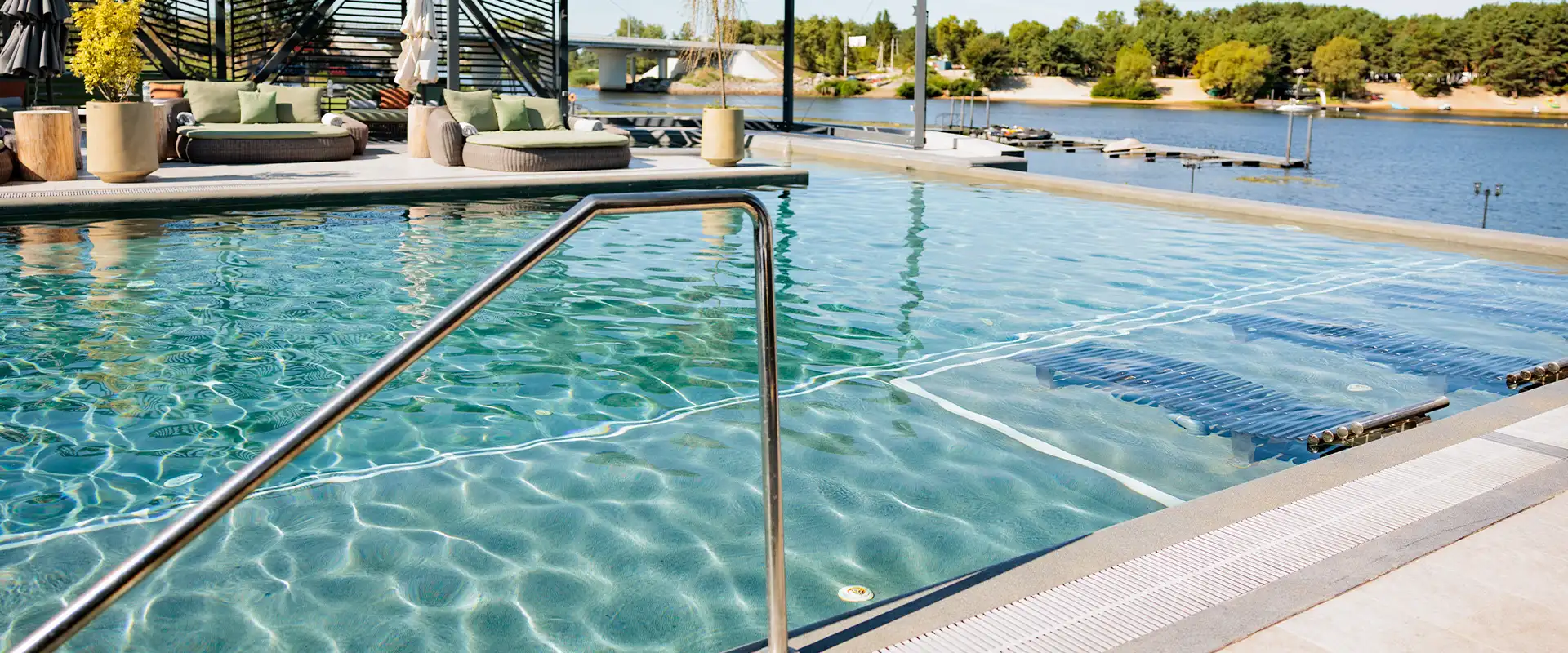A luxurious outdoor swimming pool overlooking a scenic lake, featuring modern lounge seating, glass railings, and lush green trees on a sunny day.