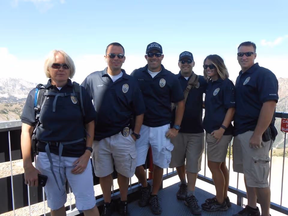 Group of six police officers in navy shirts and khaki shorts standing on a mountain overlook with a clear sky.