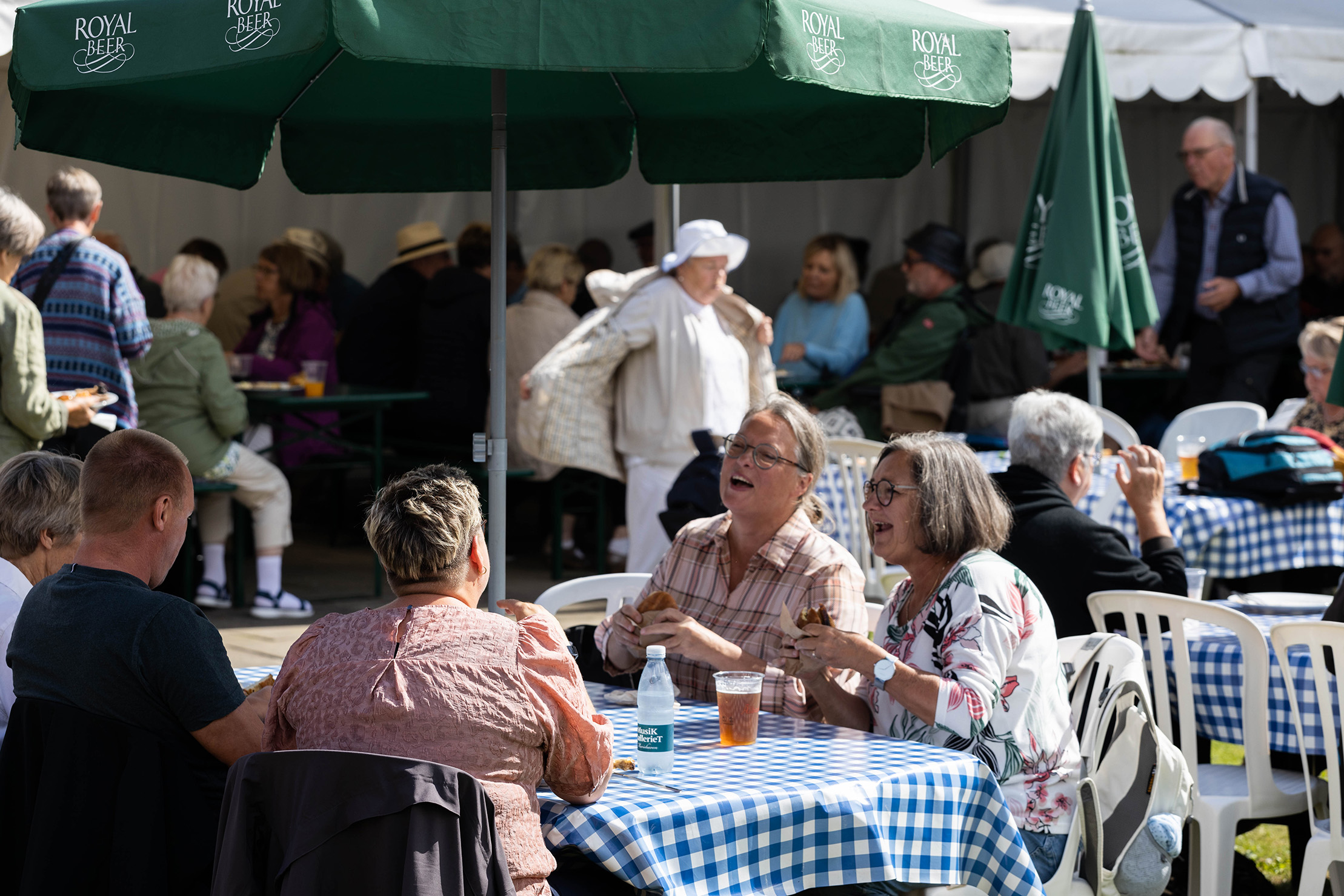 Folk sidder ved udendørs borde med blå- og hvidternede dug, spiser og snakker under grønne parasoller med Royal Beer-logo.
