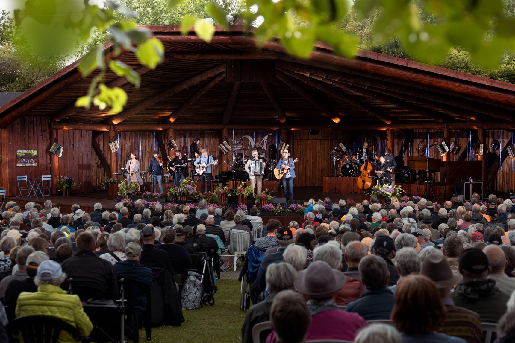 Stor gruppe musikere spiller på en oplyst træscene foran et stort publikum udendørs.
