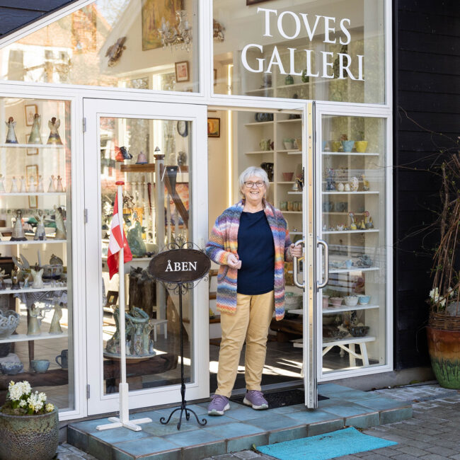 Ældre kvinde smilende i åbningen af en glasdør til Toves Galleri med hylder af keramik og kunstværker indendørs.