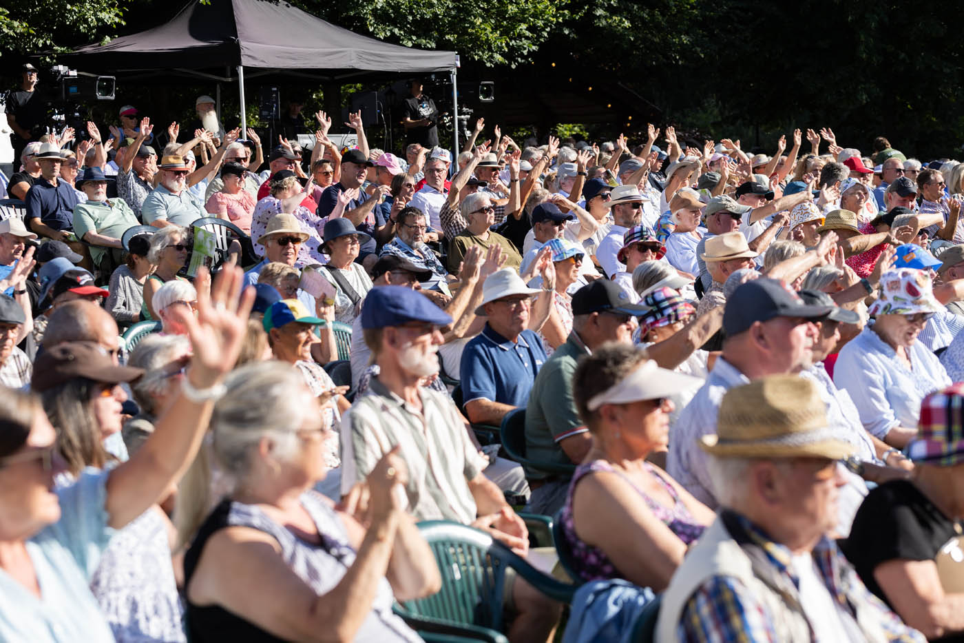 Stor gruppe ældre mennesker sidder udendørs på stolene med hænderne løftet i solen.