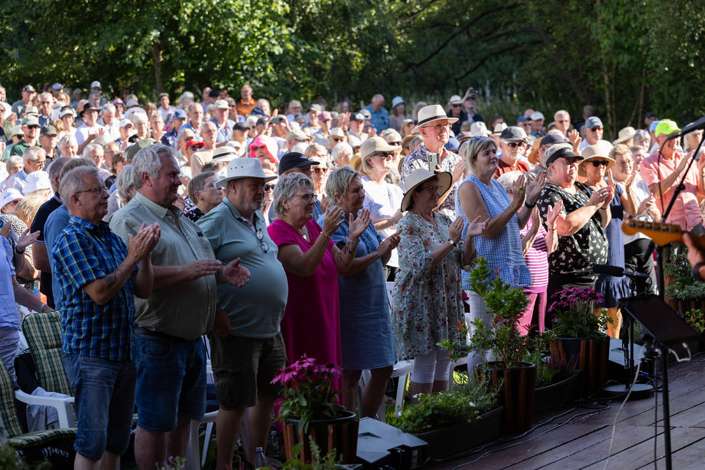 En stor flok af voksne mennesker, der klapper og står udendørs foran en scene med grønne træer i baggrunden.