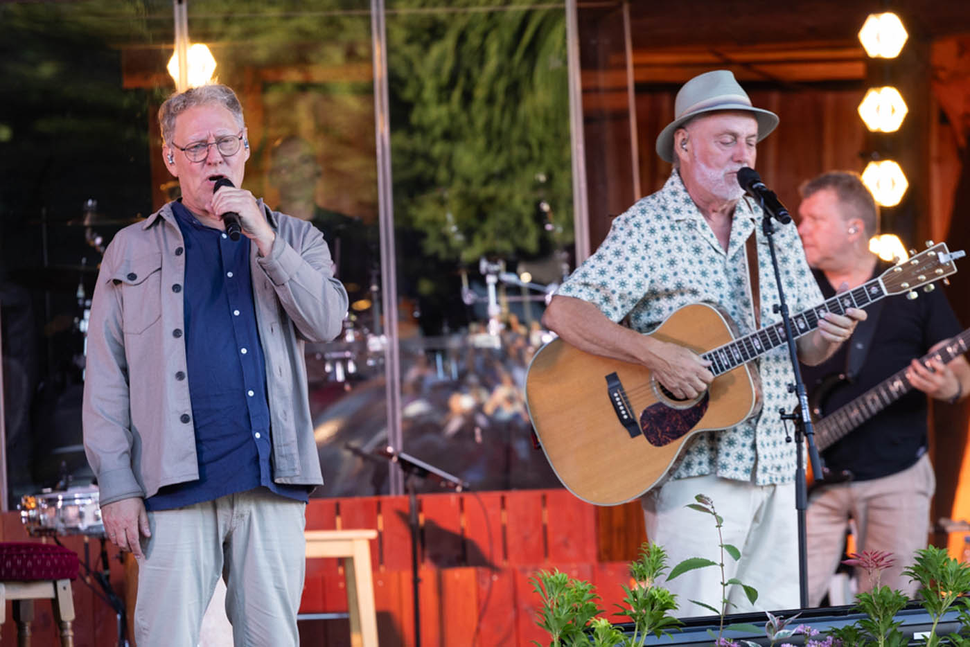 Mand og kvinde står med mikrofoner foran en flygel, mens en person spiller klaver i baggrunden.