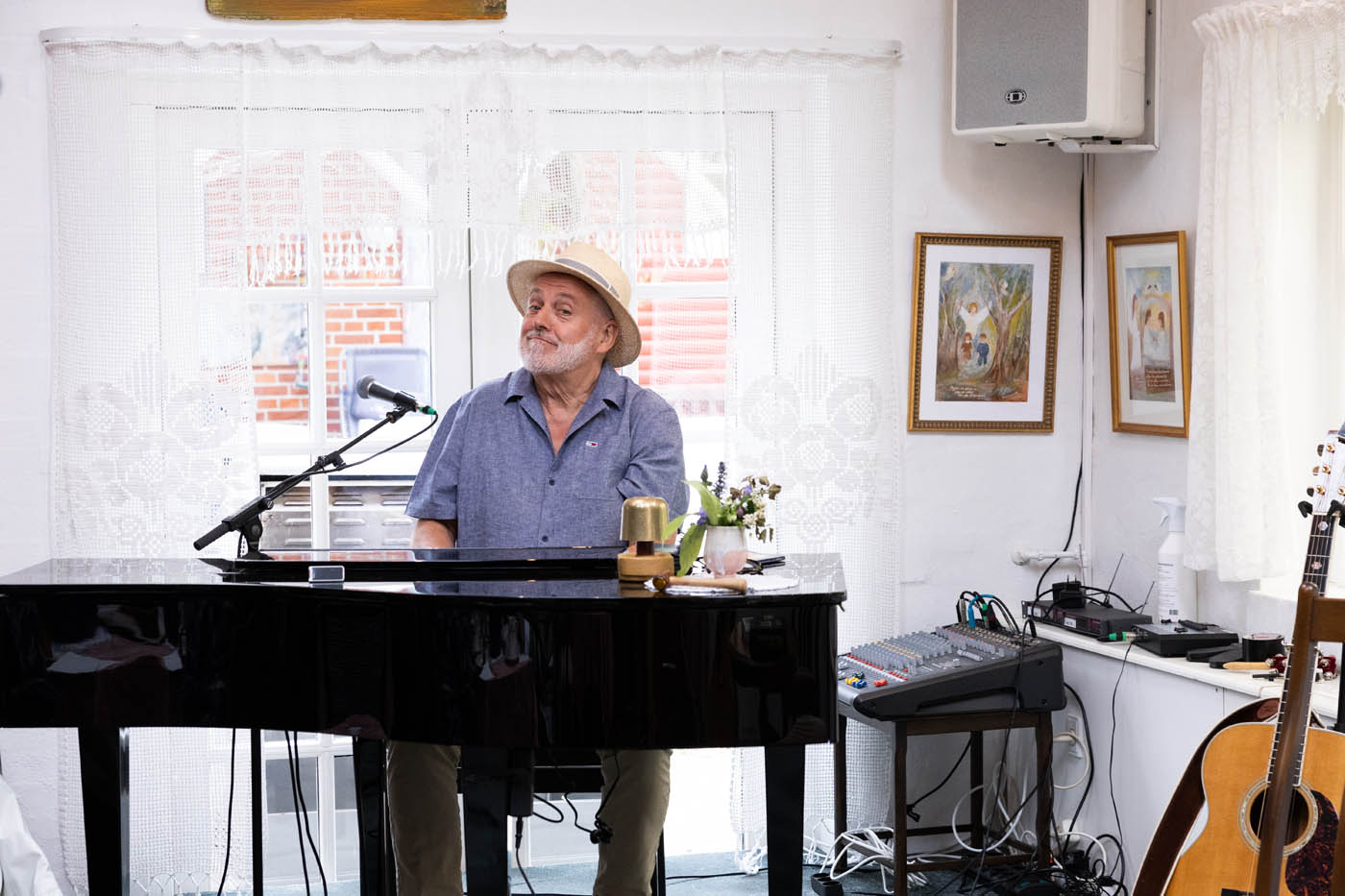 Mand i hat sidder ved et flygel med mikrofon foran, omgivet af lydudstyr og guitar i et lyst rum.