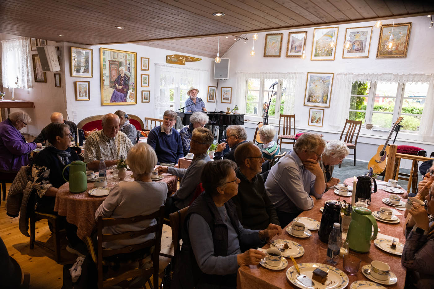 En gruppe ældre mennesker sidder omkring borde med kaffe og kage i et hyggeligt rum med malerier på væggene, mens en mand spiller klaver.