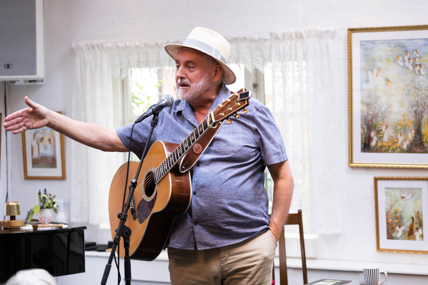 Ældre mand med hat spiller guitar og taler foran en mikrofon i et lyst rum med malerier og gardiner.