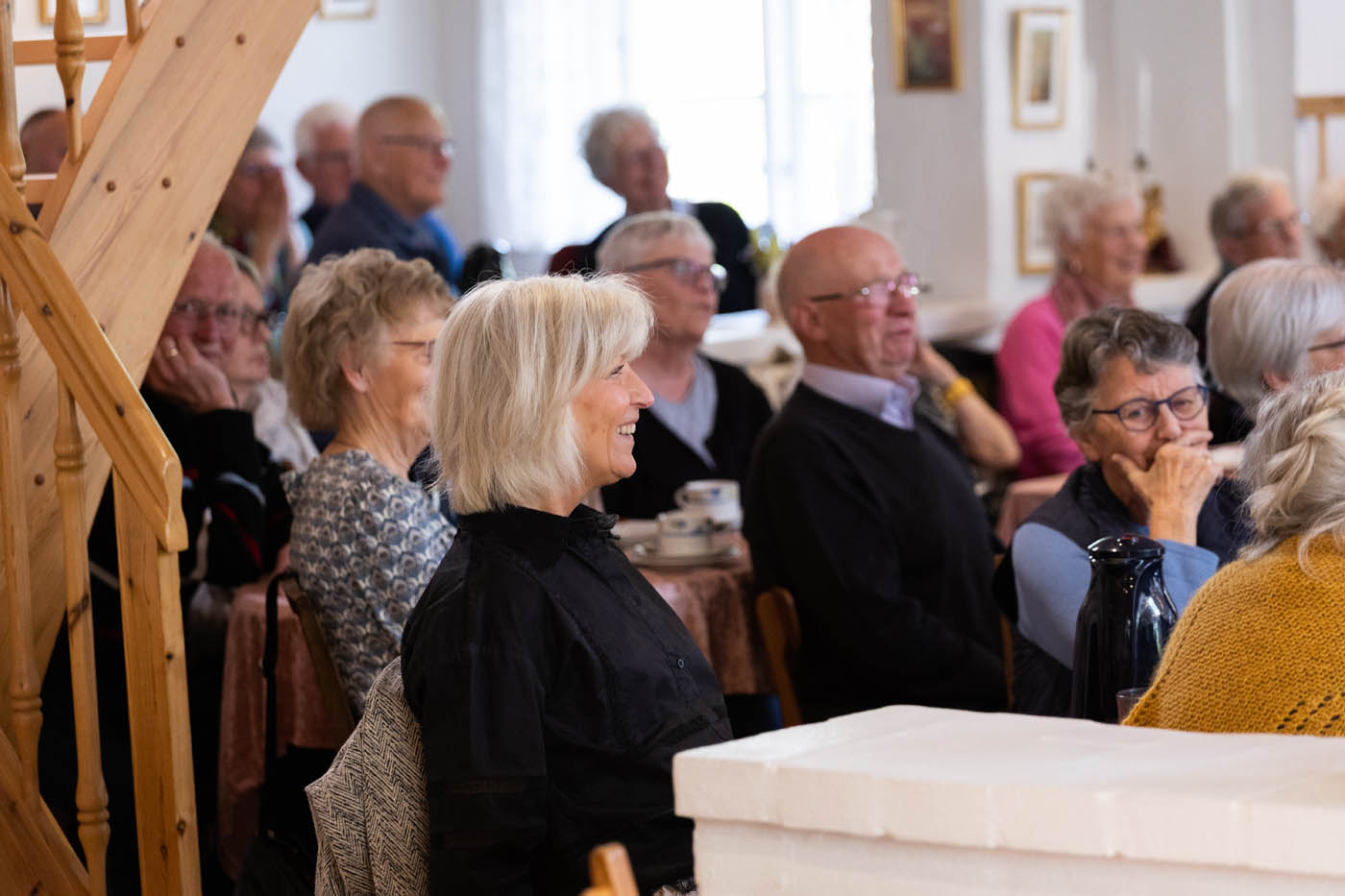 En gruppe ældre mennesker sidder samlet indendørs og lytter opmærksomt, nogle smiler.
