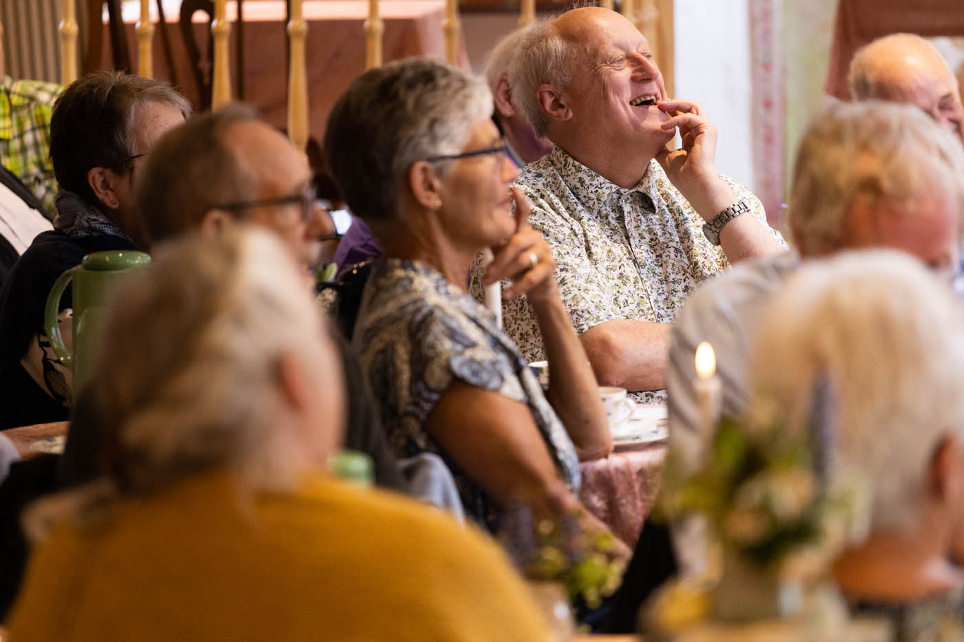 Gruppe af ældre mennesker, der sidder sammen ved et bord og lytter opmærksomt, en mand smiler bredt.