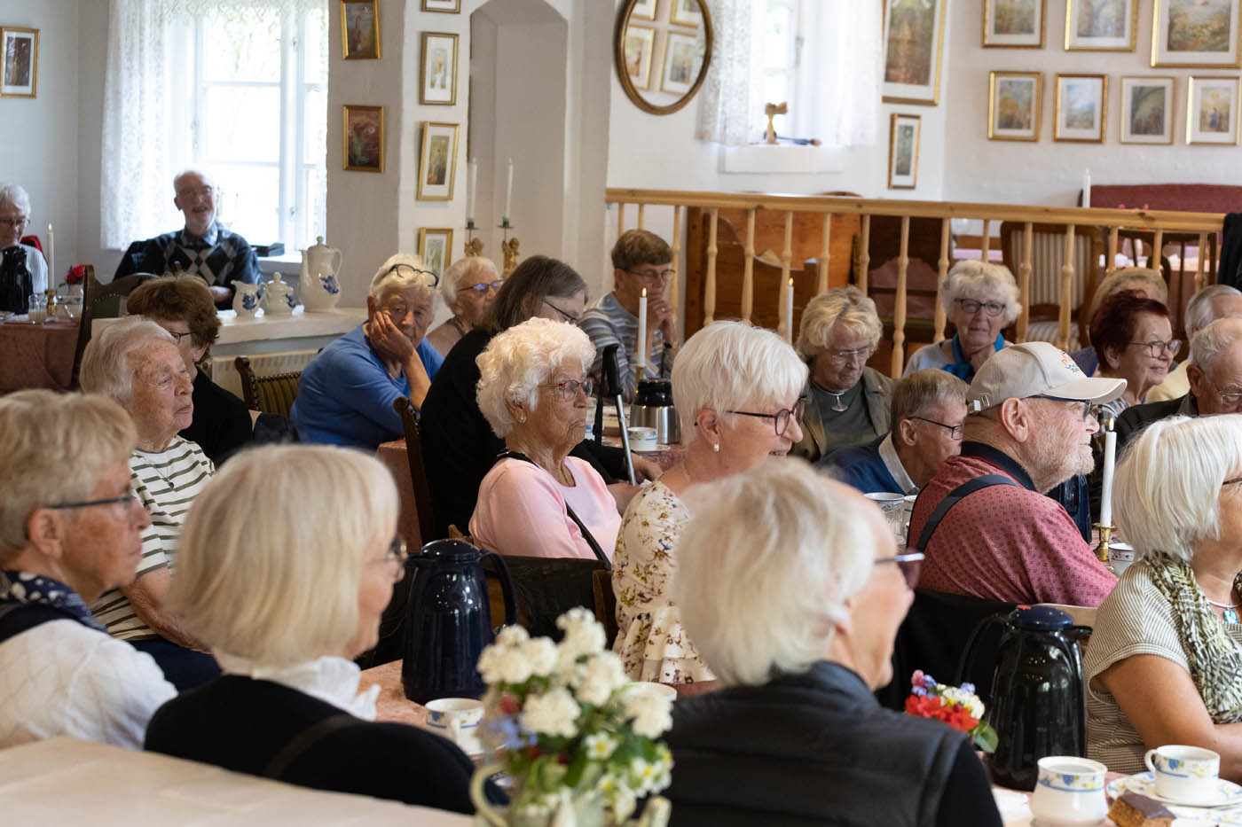 En gruppe ældre mennesker sidder samlet i et lyst rum med tekopper og blomster på bordene.