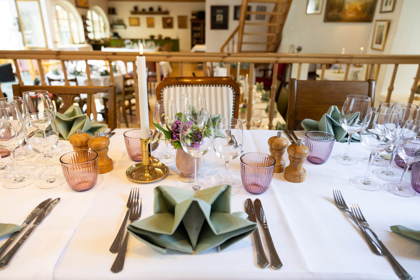 Smukt dækket middagsbord med foldede grønne servietter, vin- og vandglas, lysestage med hvidt stearinlys og blomsterdekoration.