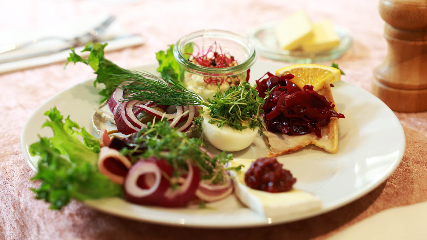 Frokosttallerken med sild, rødløg, æg med karse, rødbeder på rugbrød, ost med syltede tranebær og salatblade.