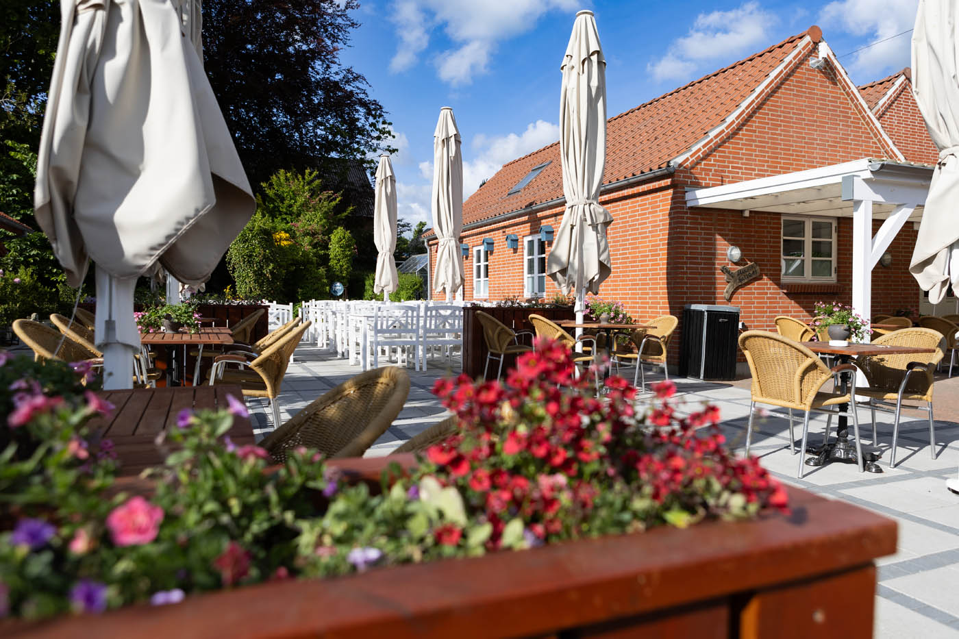 Udendørs caféområde med tomme borde og stole, lukkede parasoller og blomsterkasser foran røde murstensbygninger under en blå himmel.