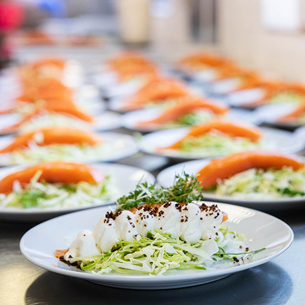 En tallerken med en gourmetret bestående af frisk kål, flødeskum med chokoladestykker og frisk timian, omgivet af flere lignende tallerkener med røget laks og salat.
