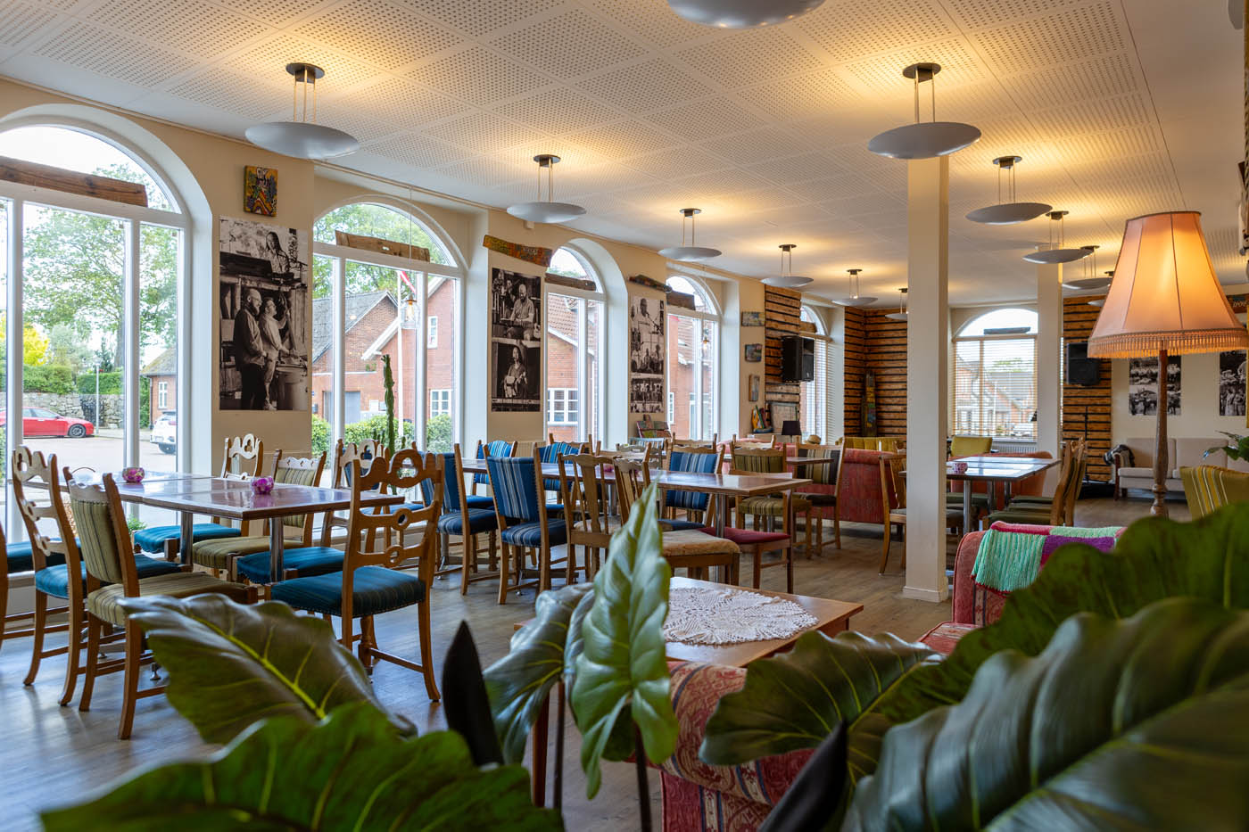 Lys café med træborde, stole med stribet polstring, store buede vinduer og bløde sofaer i forgrunden.