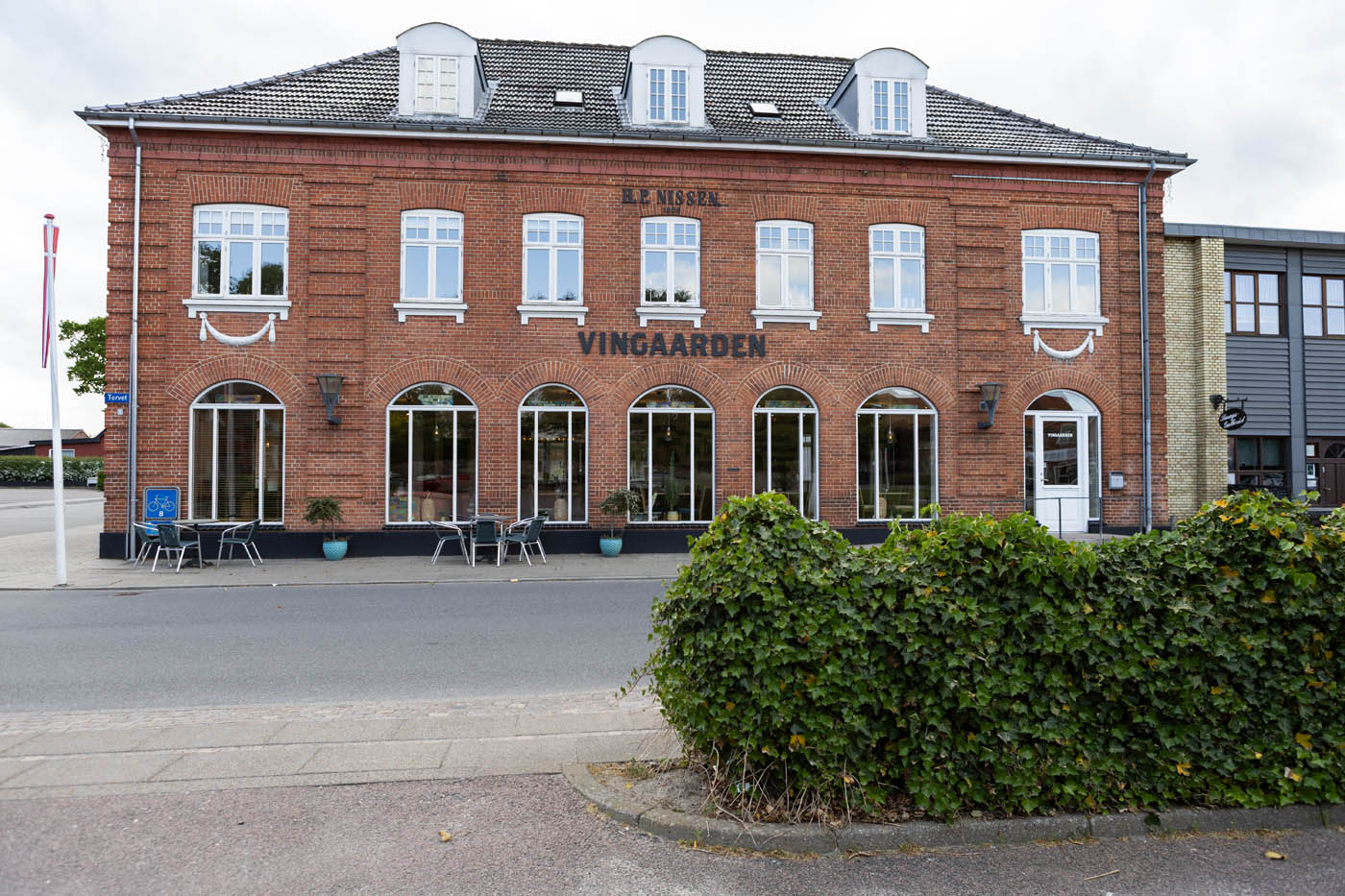 Rød murstensbygning med vinduer og skilt 'VINGAARDEN' på facaden og udendørs caféborde foran.
