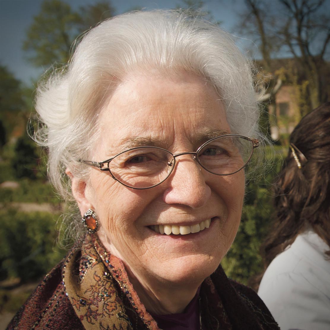 Smilende ældre kvinde med hvidt hår, briller og et smukt tørklæde udendørs i solskin.