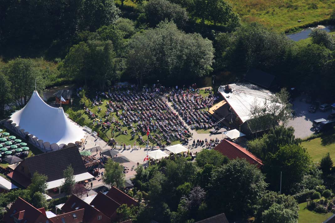 Luftfoto af et udendørs arrangement med et stort publikum siddende foran en scene og et hvidt cirkustelt i en grøn, skovklædt park.