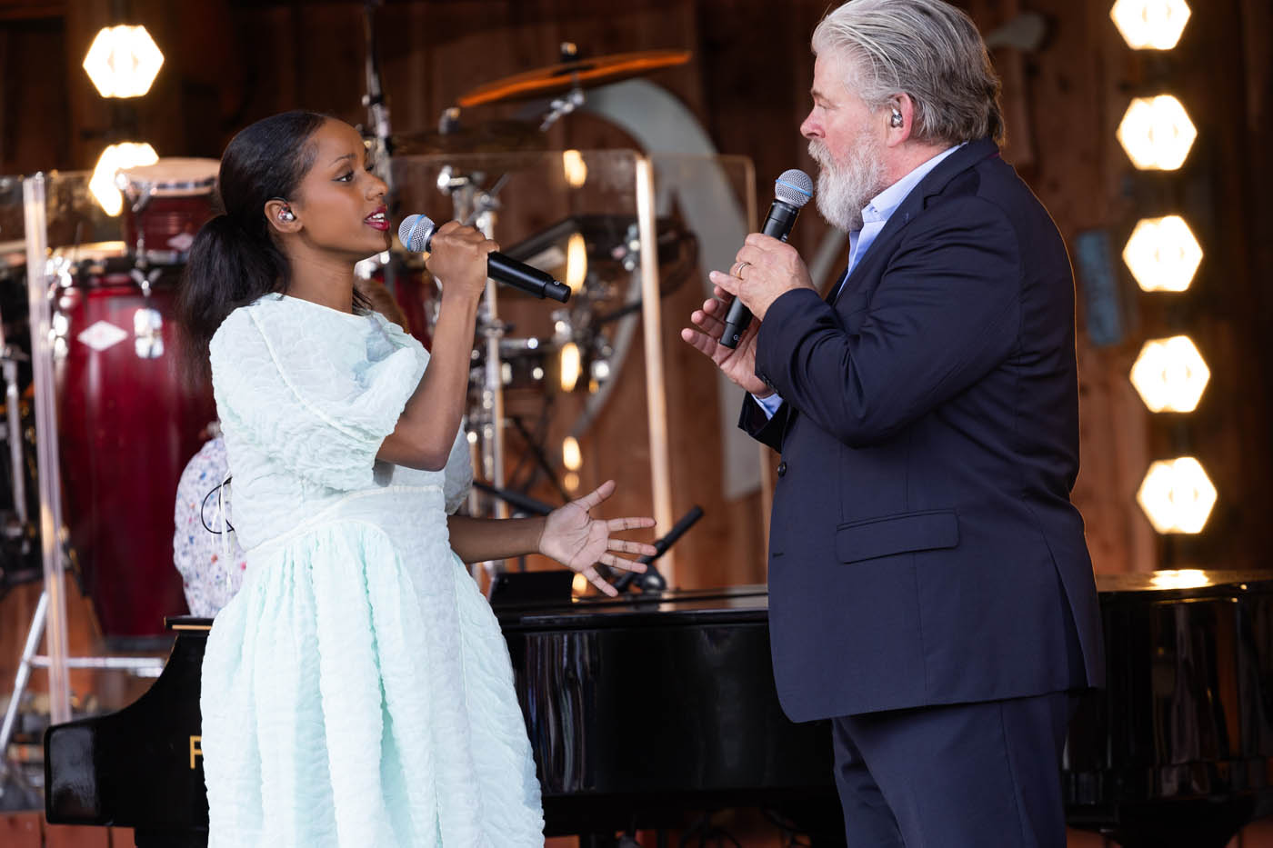 En kvinde i hvid kjole og en ældre mand i mørk jakkesæt synger sammen med mikrofoner foran et klaver på en scene.