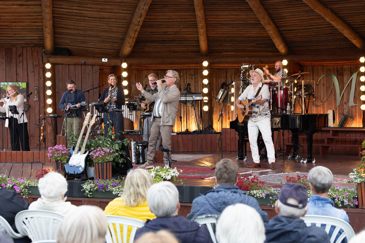 Musikgruppe spiller live på en træscene med publikum siddende på hvide stole foran scenen.