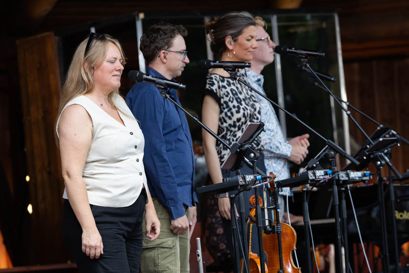 Fire personer står på række ved mikrofoner, klar til at synge eller optræde på en scene med musikudstyr og en violin.
