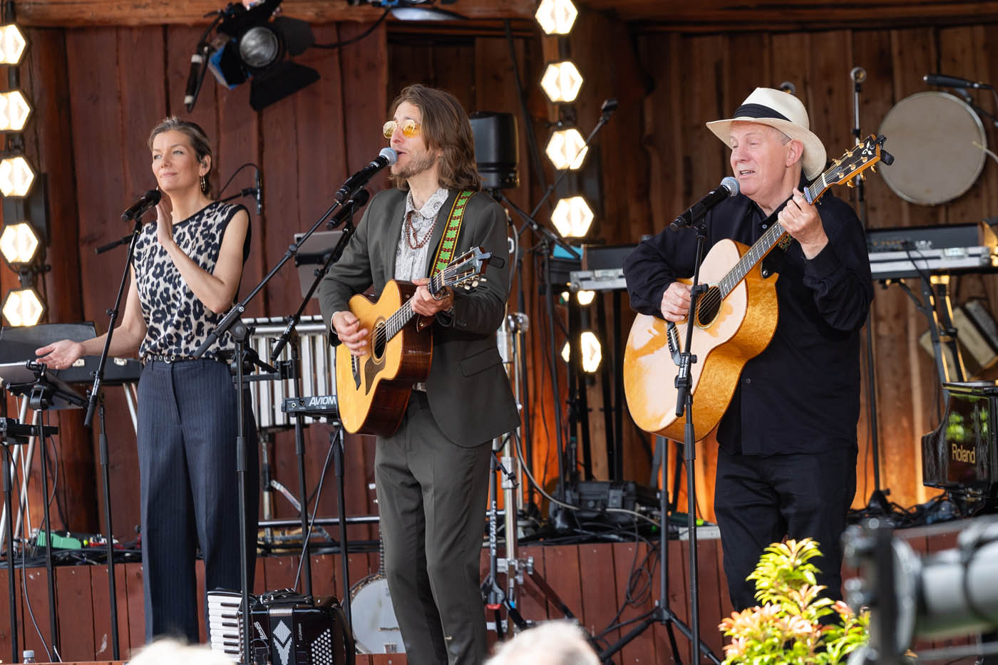 Tre musikere, to mænd med guitarer og en kvinde med mikrofon, optræder på en scene med træbaggrund og lysinstallationer.