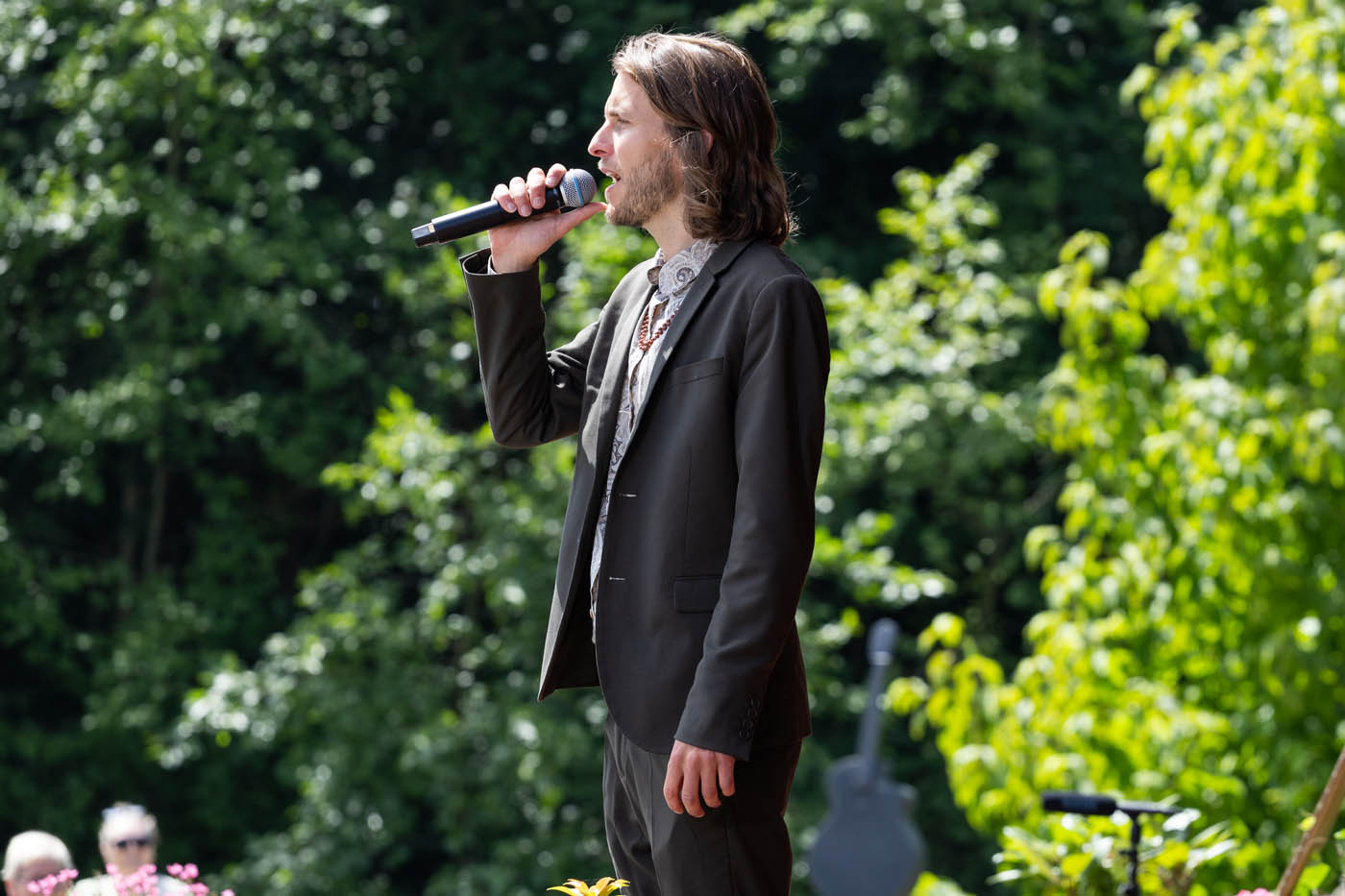 Mand med langt hår iført jakkesæt, der synger i mikrofon udendørs med grønne træer i baggrunden.