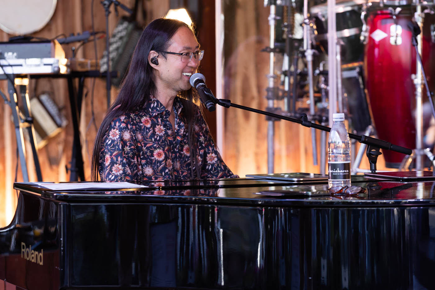 Mand med langt hår sidder ved et Roland-flygel og synger i en mikrofon under en musikoptræden.