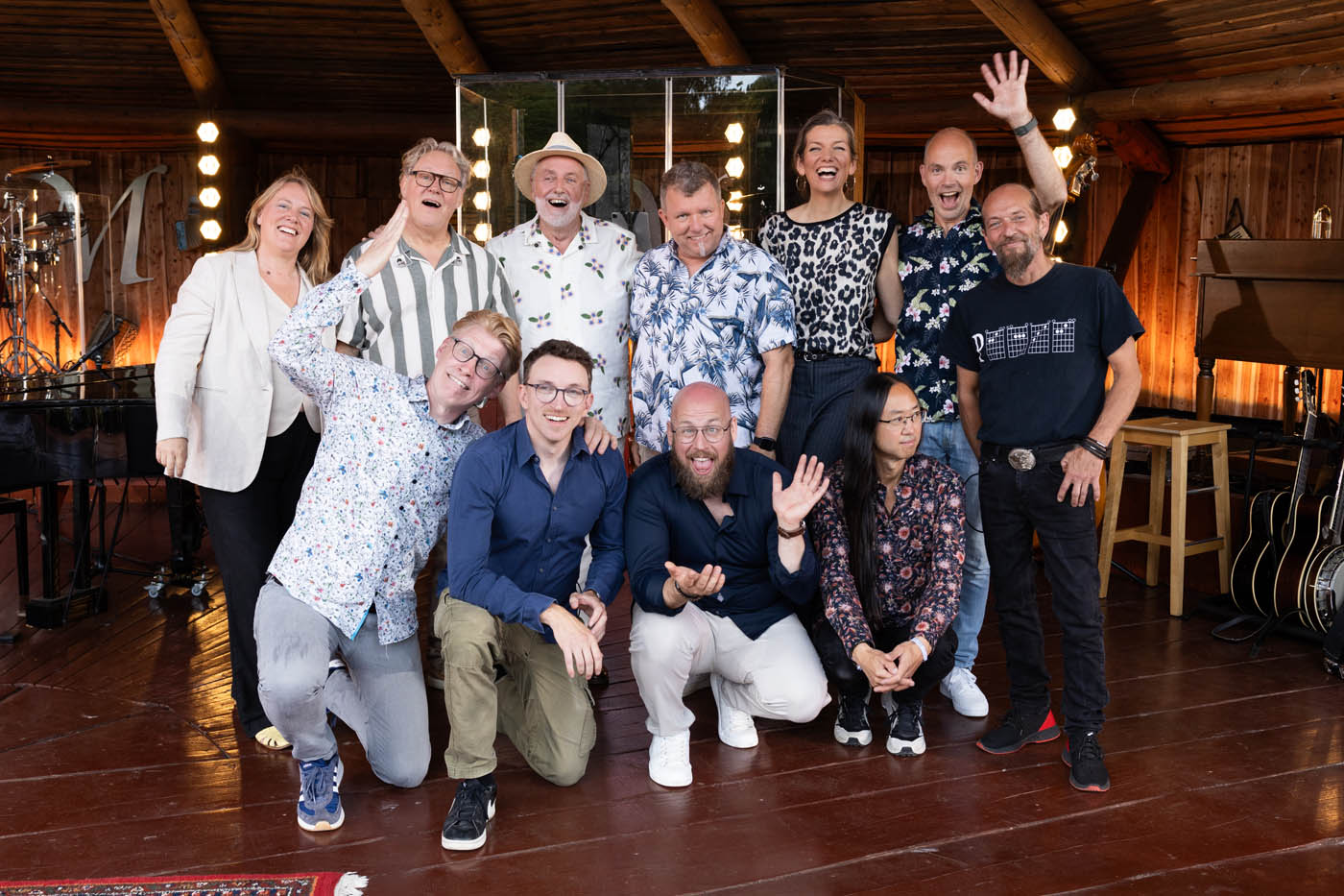 En gruppe på 11 glade voksne i afslappet tøj poserer i et træbeklædt rum med musikinstrumenter.