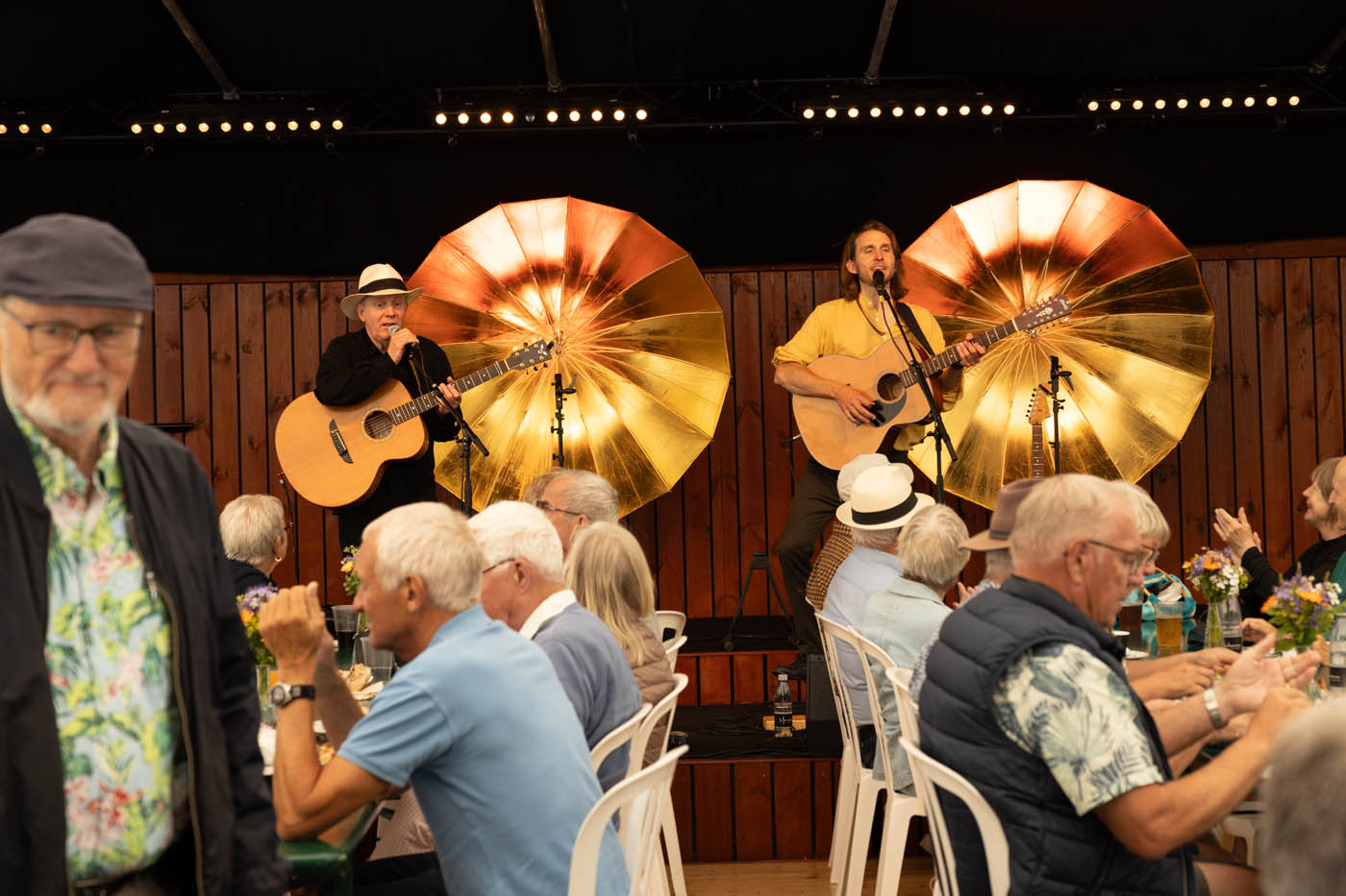 To musikere spiller akustisk guitar og synger foran et træpanel med store, gyldne reflektorer, mens ældre mennesker sidder ved borde og lytter til musikken.