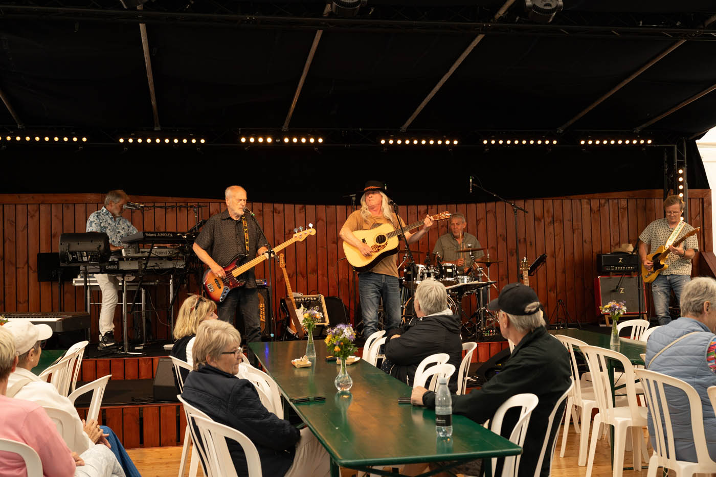 Et band med fem musikere spiller på en scene foran et publikum, der sidder ved grønne borde med blomster i vaser.